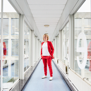 Stephanie Wehner in a red suit standing in a glass-paneled corridor at Delft University of Technology in the Netherlands. Her reflection appears in the glass to the right and left.
