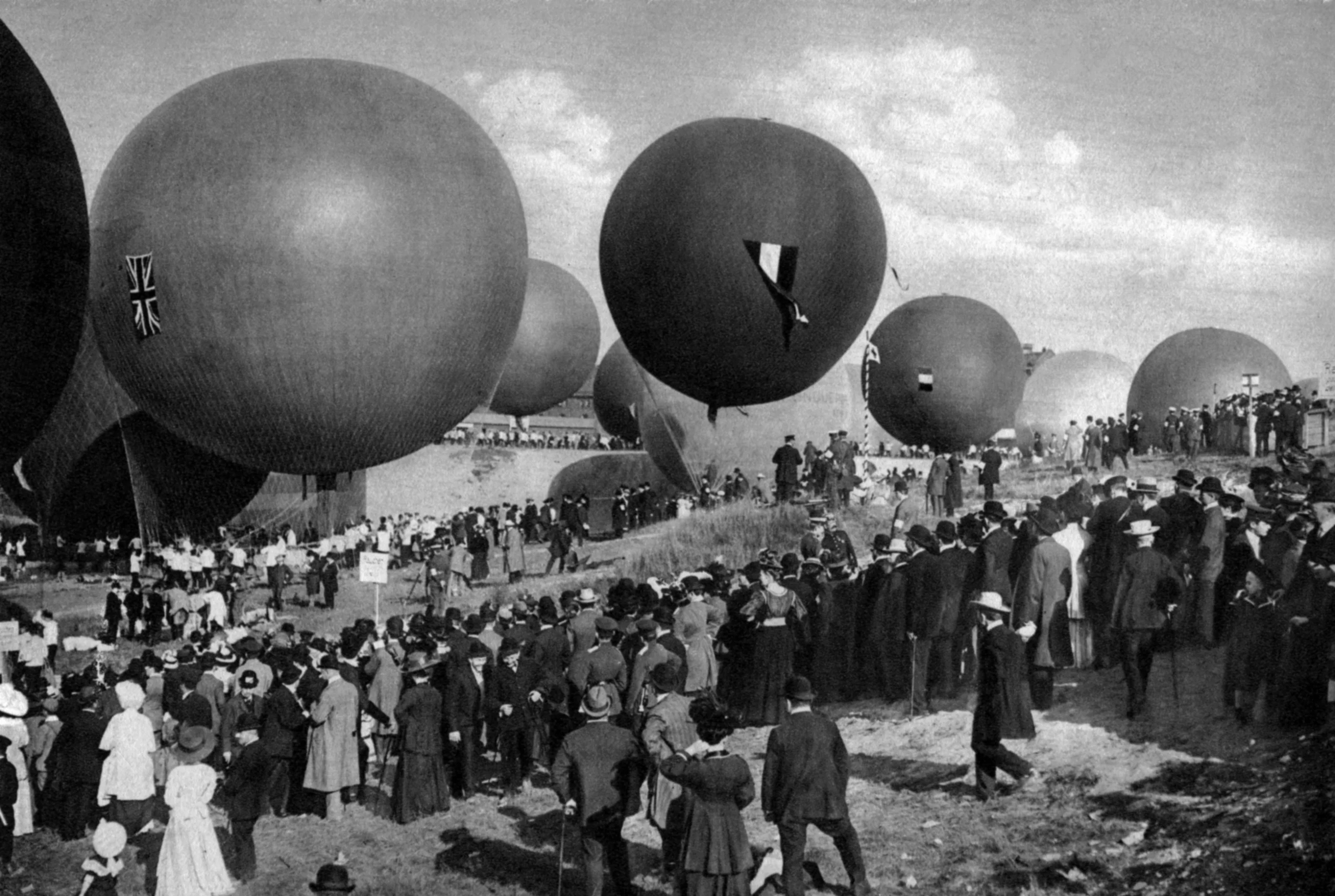 Black-and-white photograph of gas balloons getting ready to launch in front of a crowd of people.
