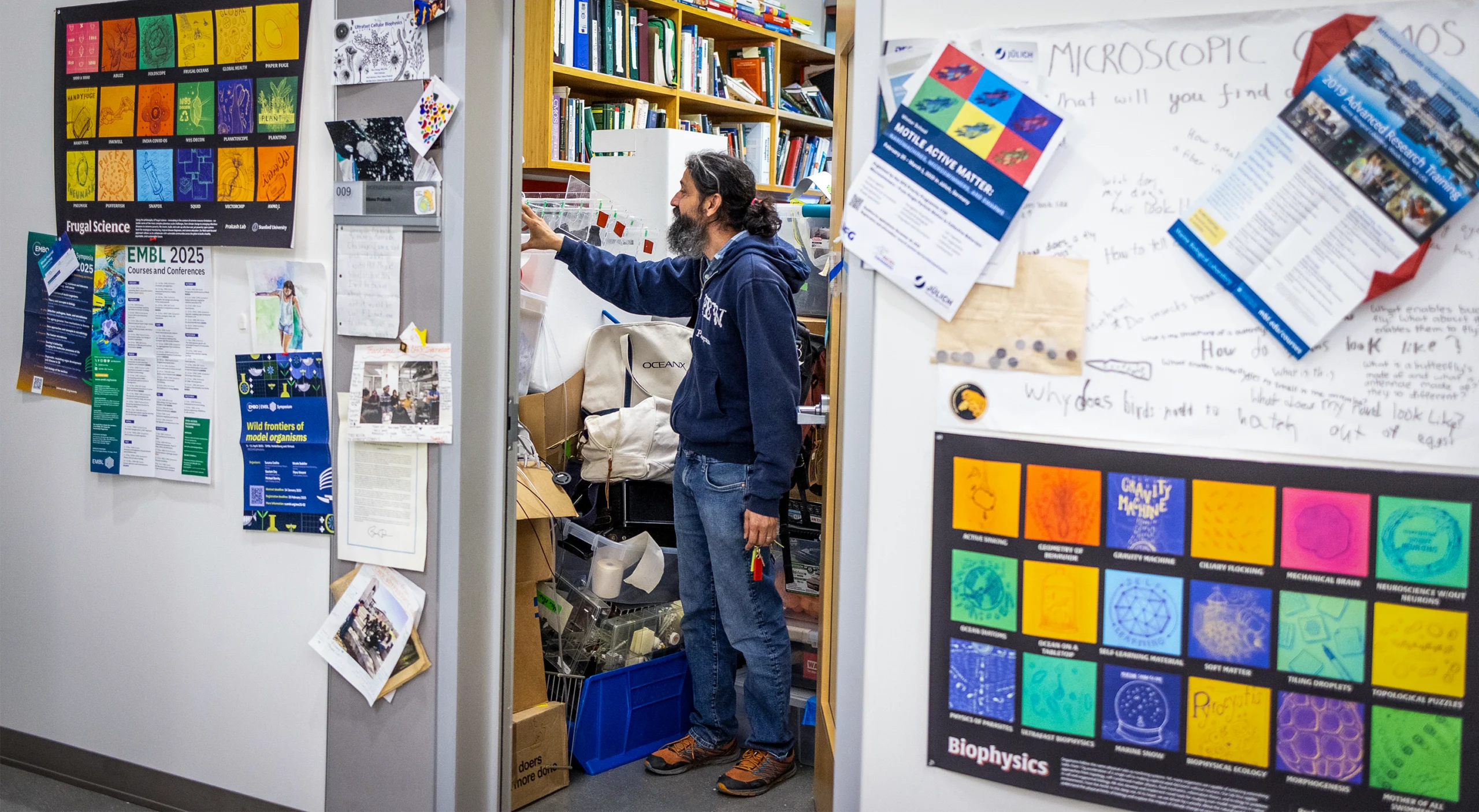 Manu Prakash stands in his office; on either side of the door are posters for “frugal science” and “biophysics.”
