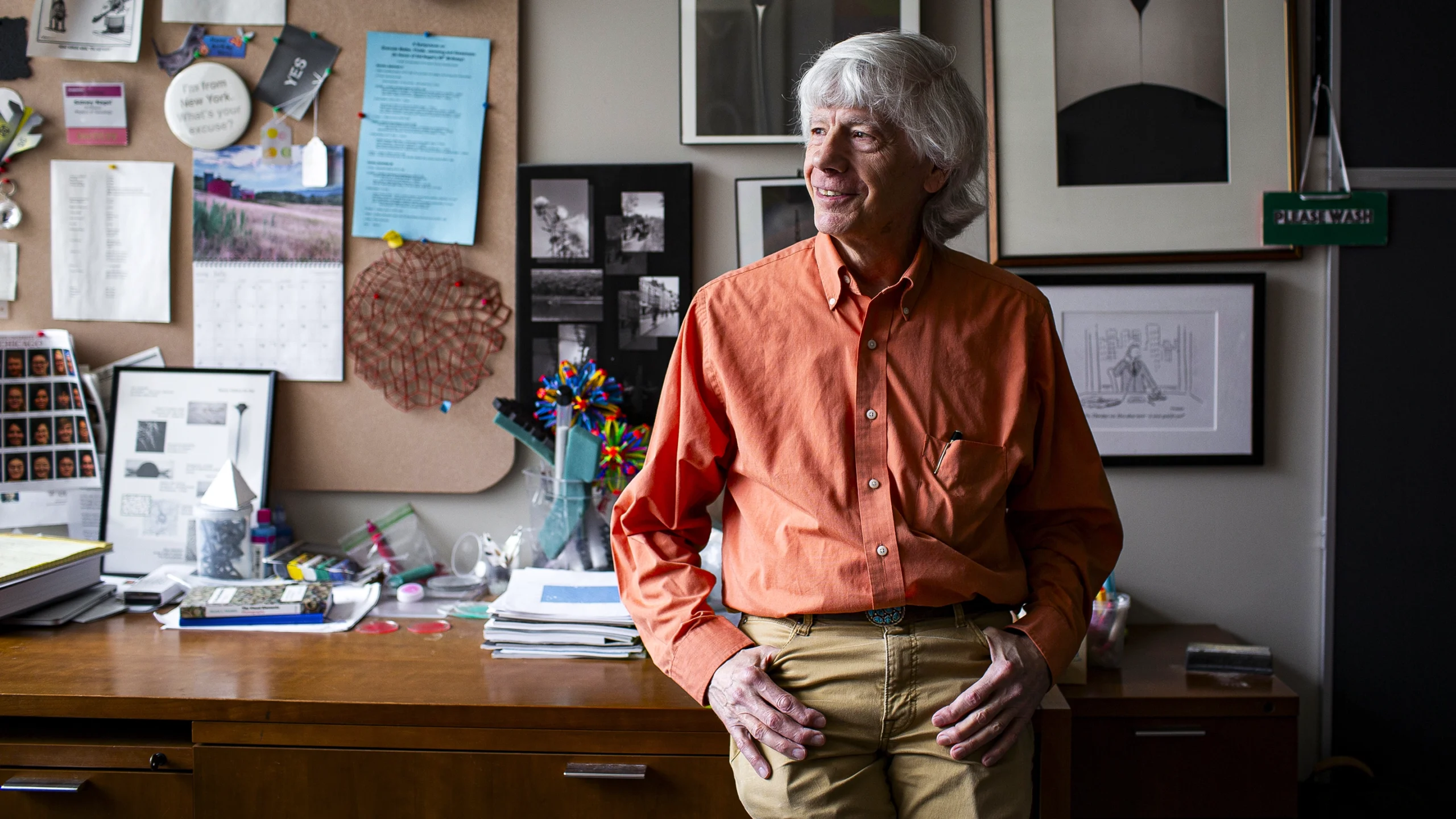 A man in an orange shirt leans against his desk, with research photographs hung on the wall behind him.