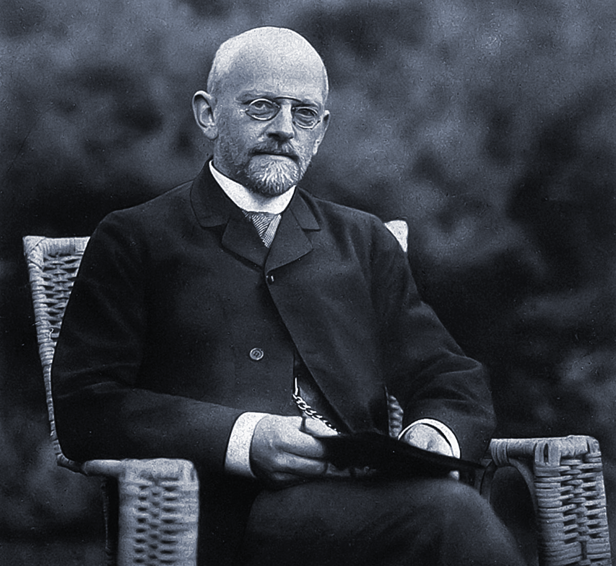 Black-and-white photo of a bald man sitting in a wicker chair.