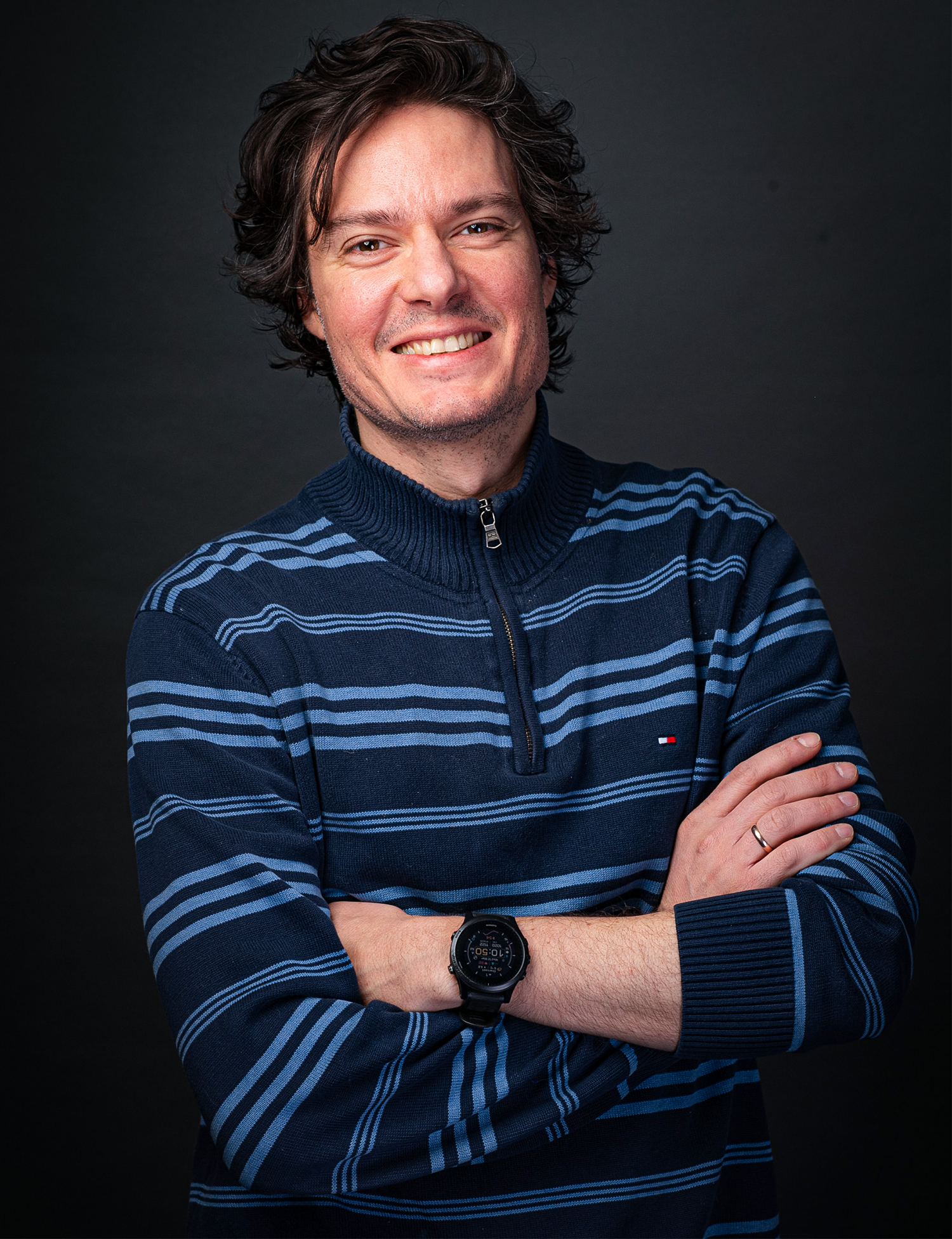 A smiling man stands with his arms crossed in front of a dark background.