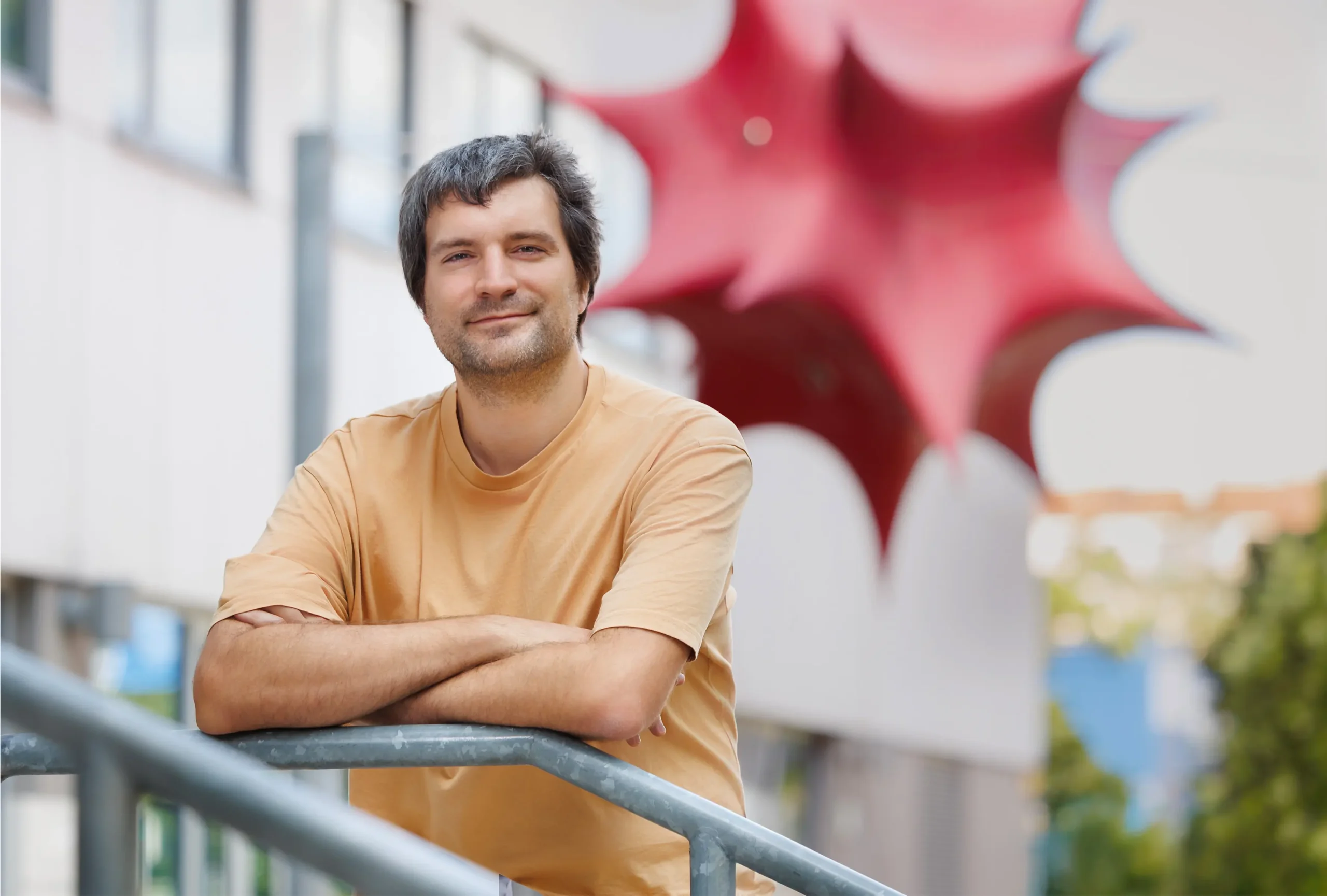 Man in orange T-shirt standing outside in front of a red geometric sculpture.