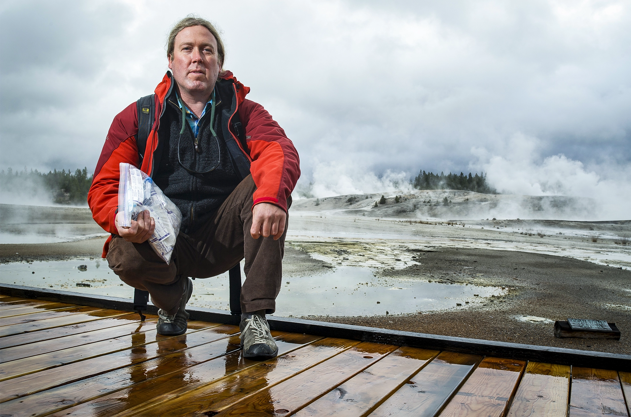 Eric Boyd stands on a boardwalk holding a bag of microbial samples collected from the hot spring behind him.
