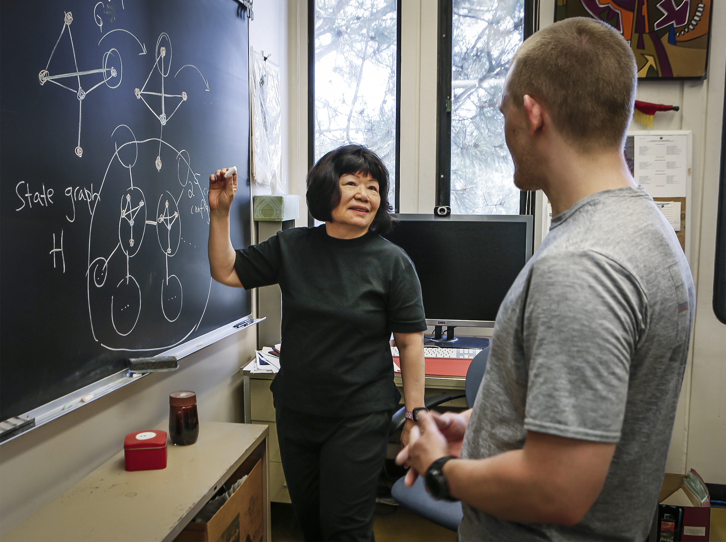 Woman in black clothes and man in gray T-shirt standing in front of a blackboard.