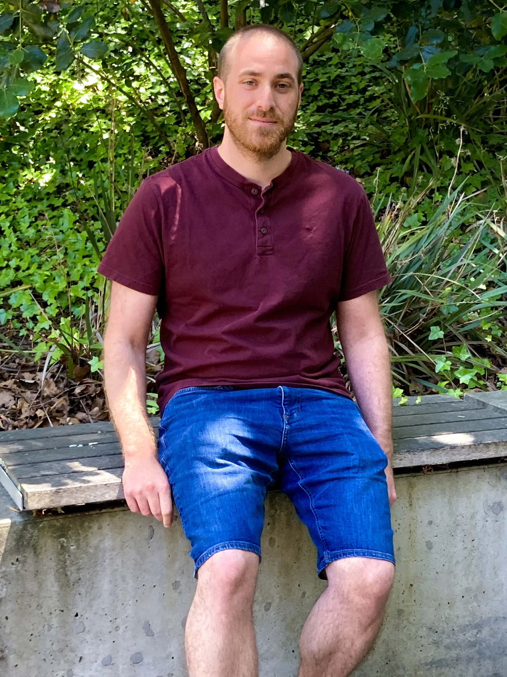 Gal Arnon, a computer scientist, is wearing a purple T-shirt and blue denim shorts, and is sitting on a wooden bench with plants in the background.