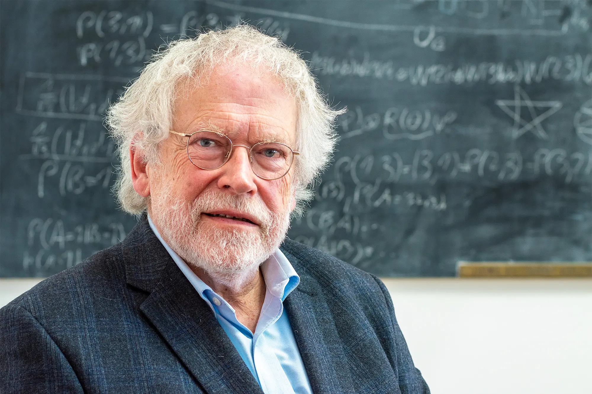 A white-haired and white-bearded man sits in front of a blackboard.