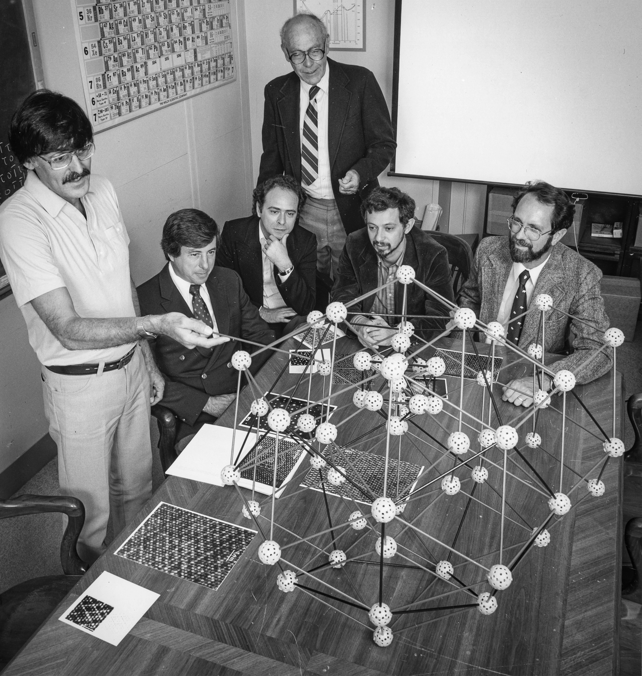A man with a mustache points at a complicated ball-and-stick model of a quasicrystal as four men look on.