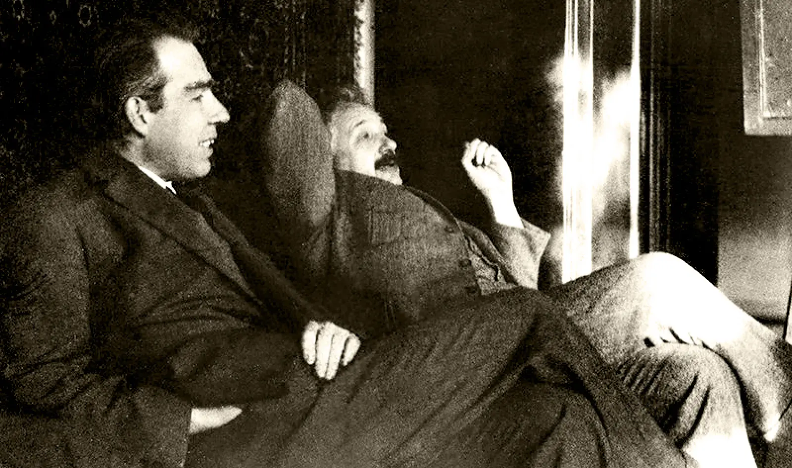 : A black-and-white photo of two men wearing suits and sitting side by side, leaning back, while having a discussion.