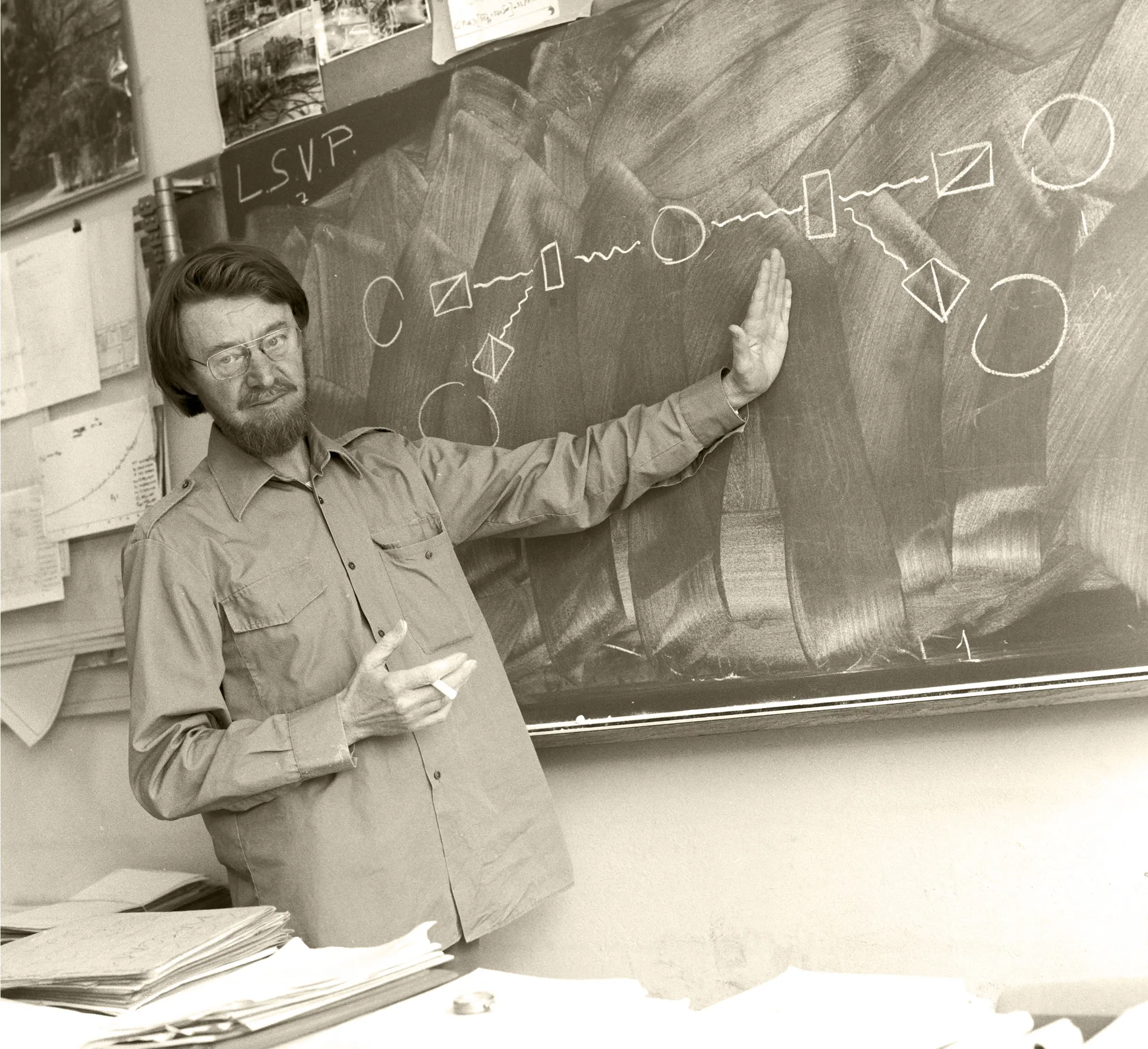 A bearded man gestures at a chalkboard.
