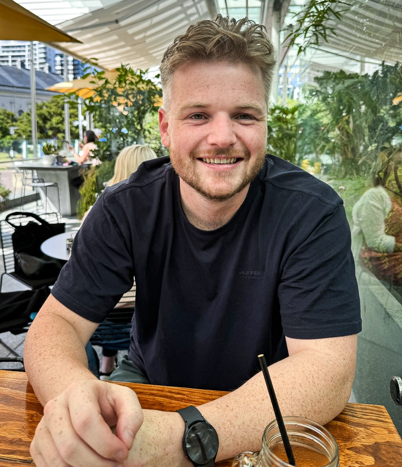 Jordan Douglas in a black T-shirt sits at a table in an outdoor café with tables and plants in the background.