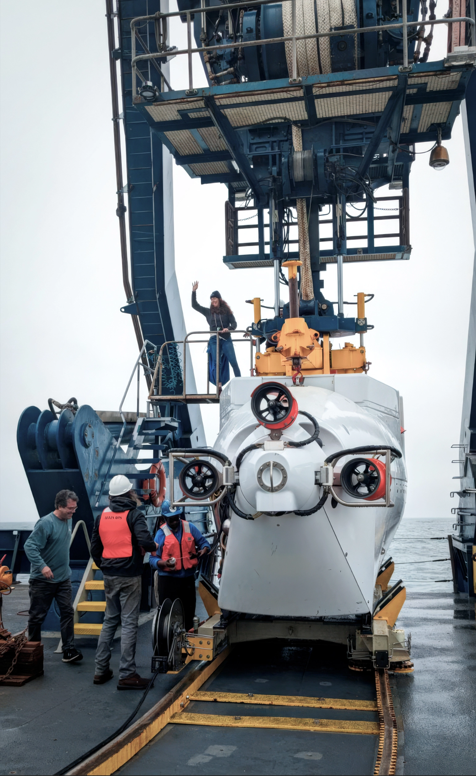 Lloyd, waving her hand, stands on the deck of the submersible Alvin, which is itself docked on the deck of a ship