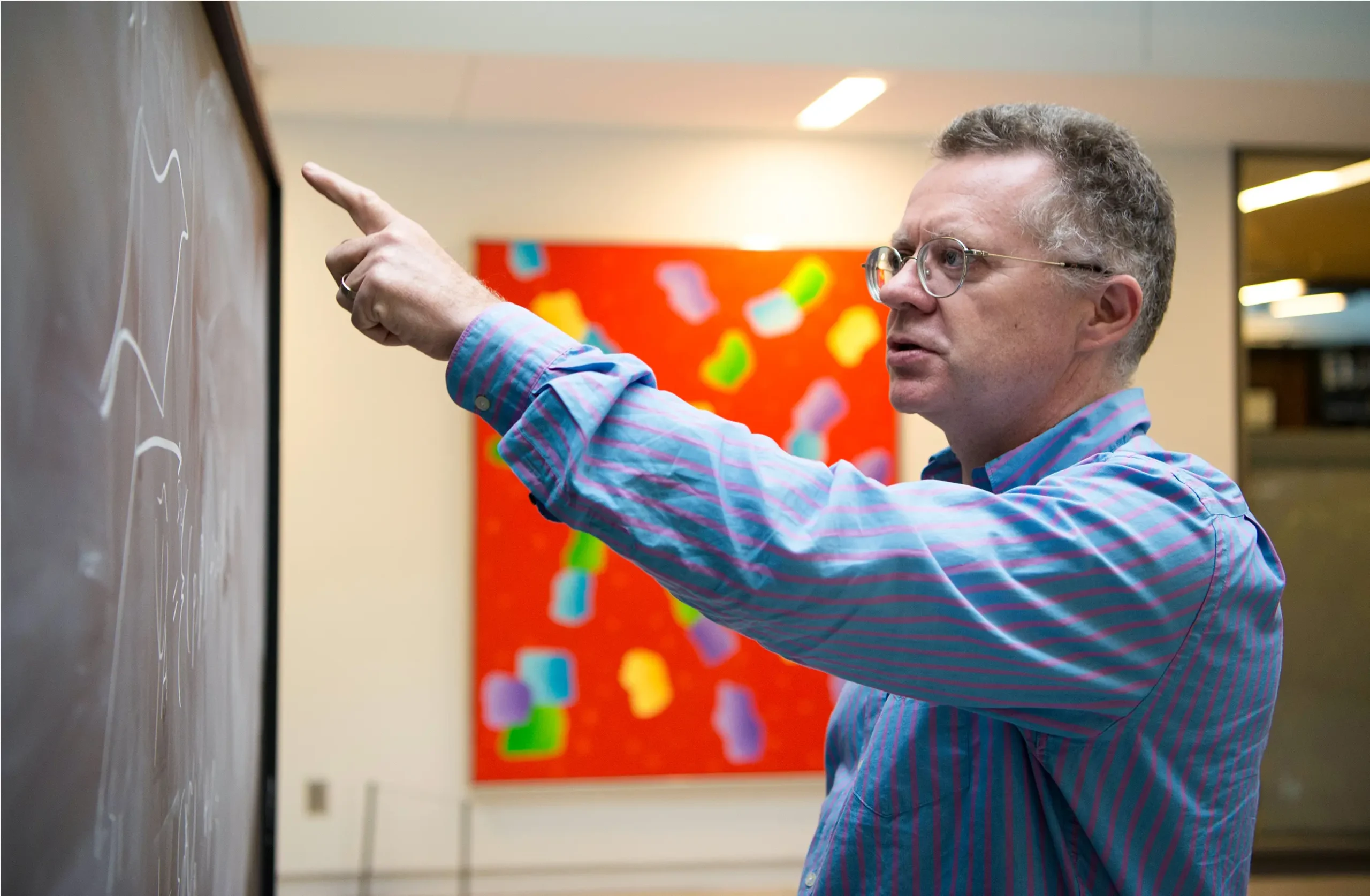A man wearing a blue shirt with red stripes points at a blackboard.