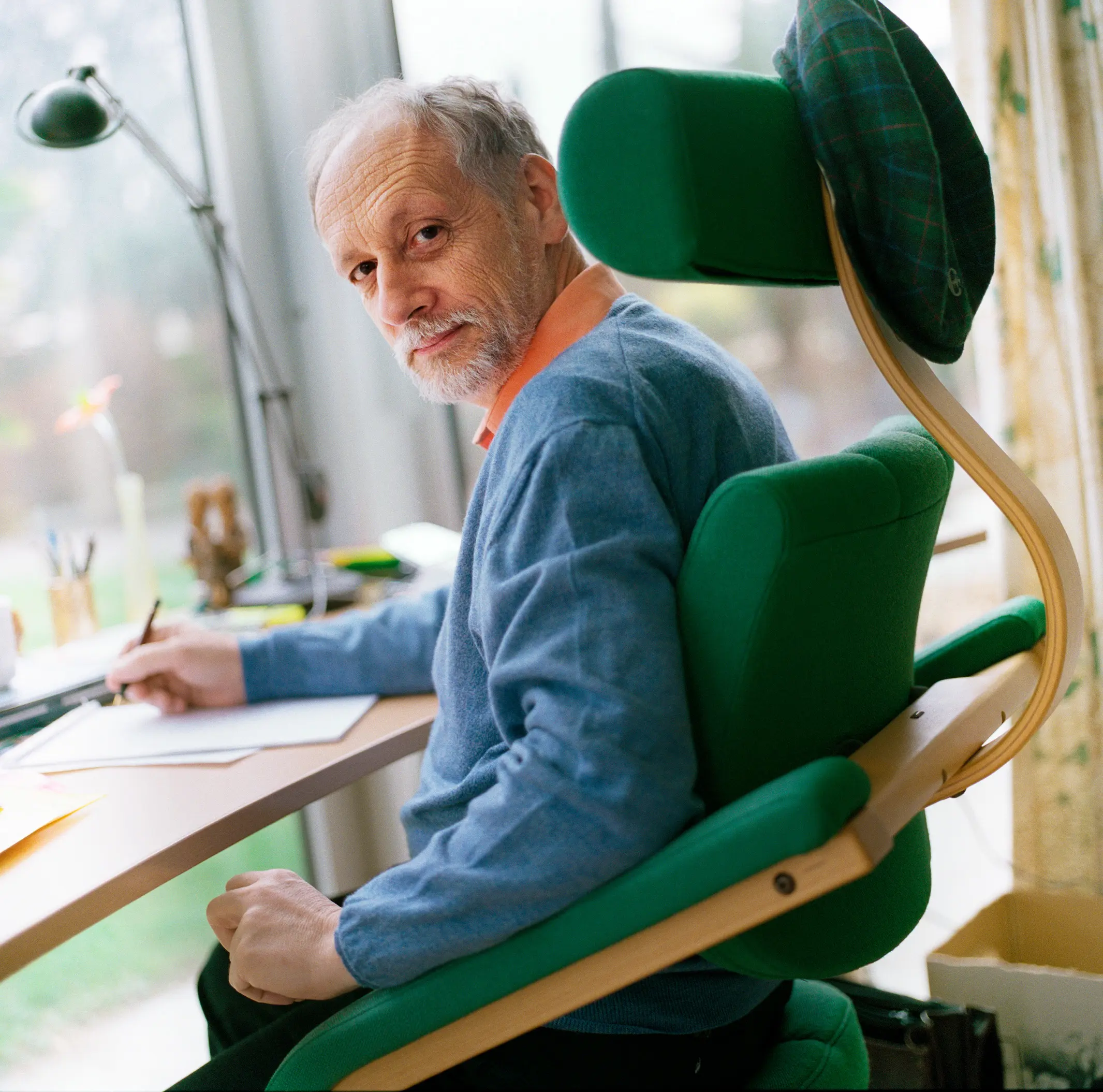 A man wearing a blue sweater sits in a green chair at a desk in a sun-drenched room.