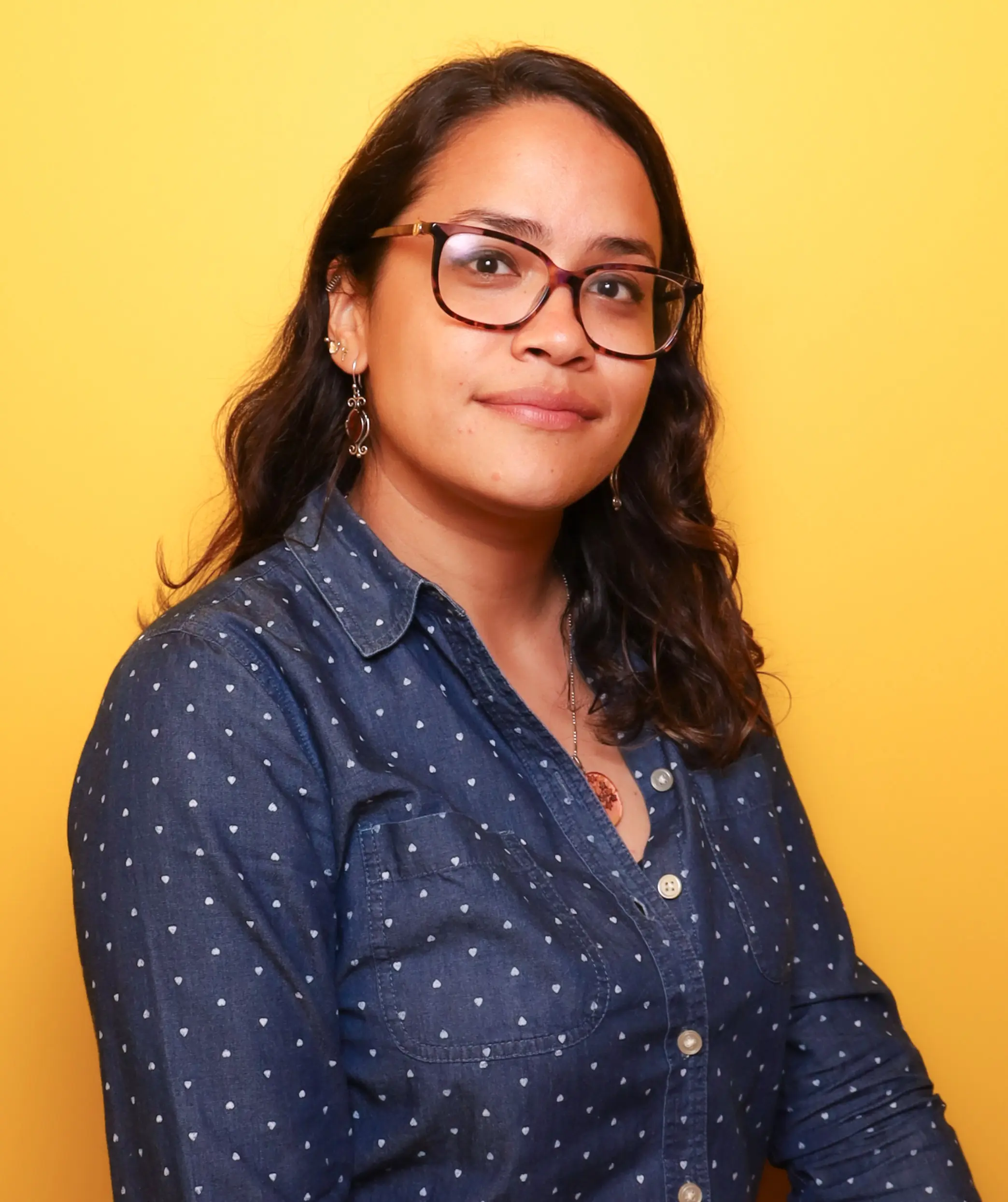 A woman in a polka-dot shirt and glasses standing against a yellow background.