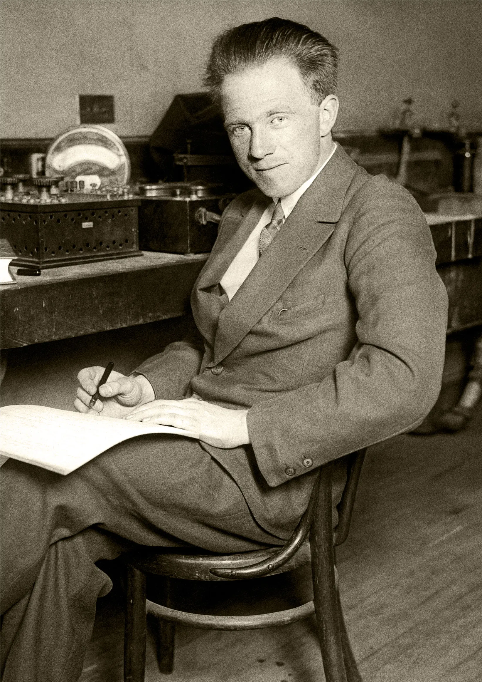 Sepia photo of a young man with a quiet smile writing on a notepad on his lap.