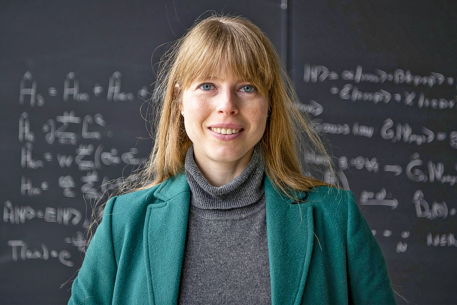 A woman in a green jacket stands in front of a blackboard.