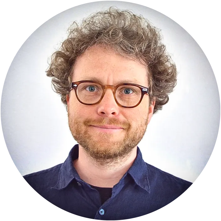 A smiling Florian Wollweber looks directly into the camera. He has short curly fair hair and a fair beard, and he is wearing wide oval-rimmed glasses, and a navy blue shirt.