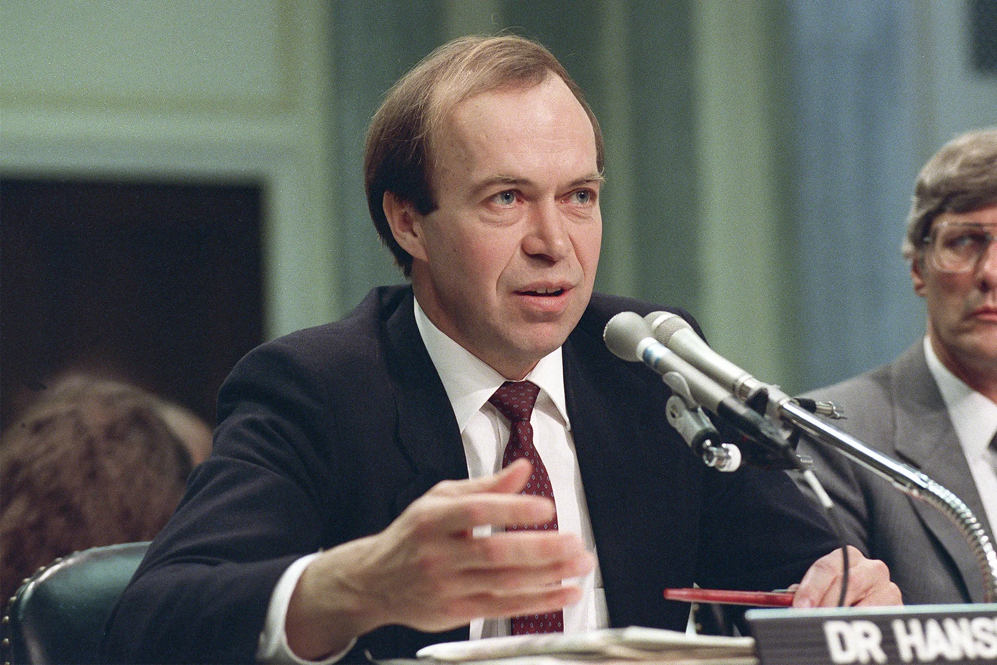 Photo of a seated man in a suit speaking into a microphone.