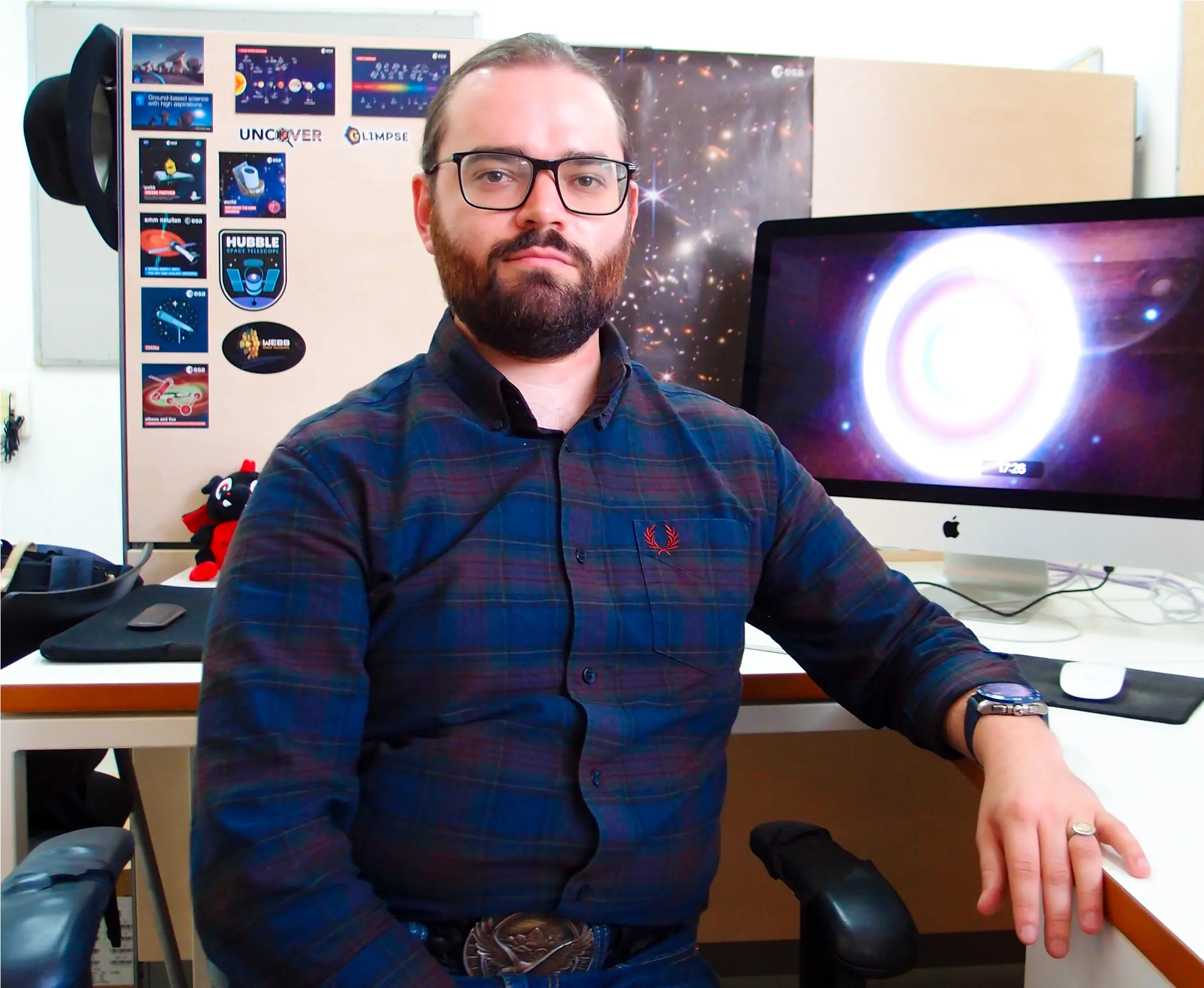 A bearded man wearing a dark blue shirt sits in front of his desk.