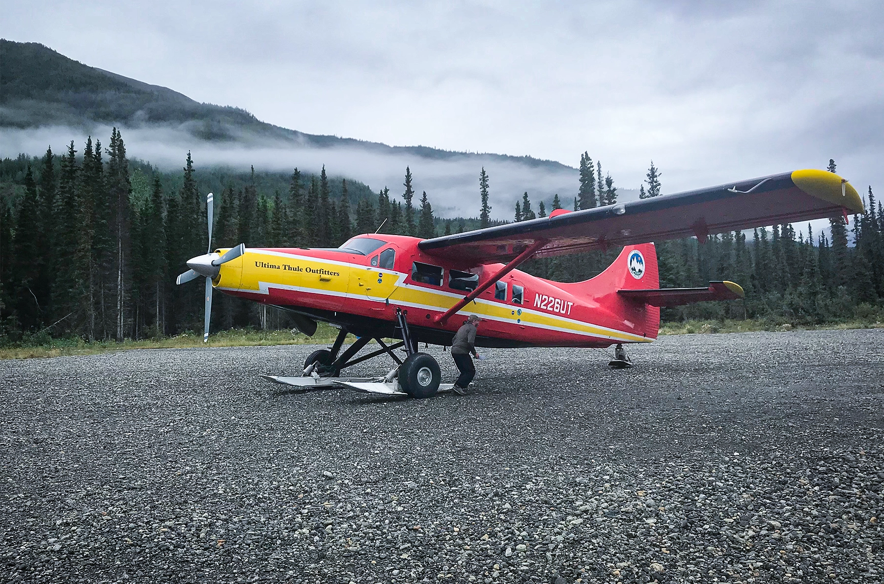 A small red-and-yellow plane sits in front of trees and a mountain.