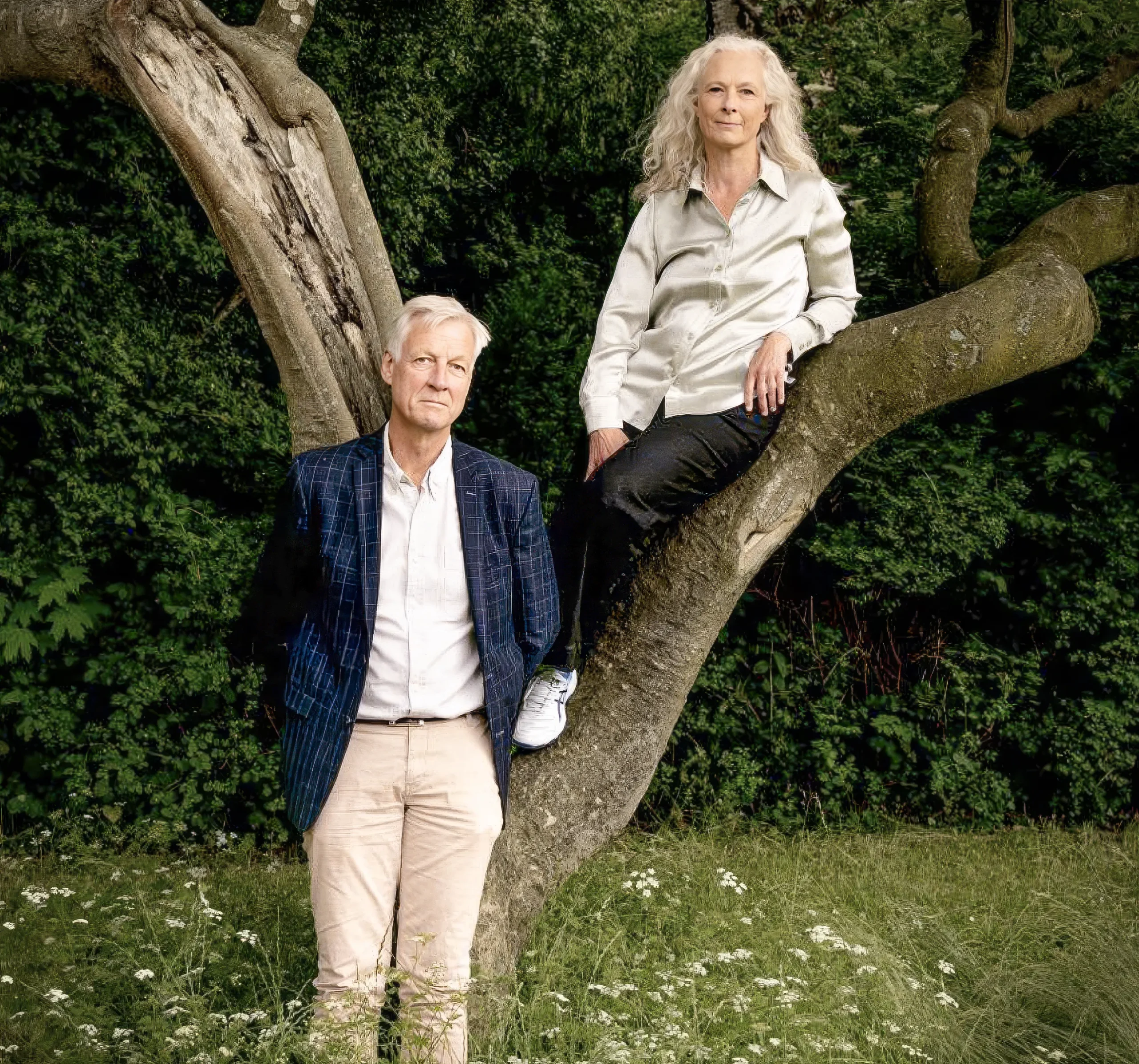 A white-haired man standing in front of a tree, and a white-haired woman sitting in one of its branches.