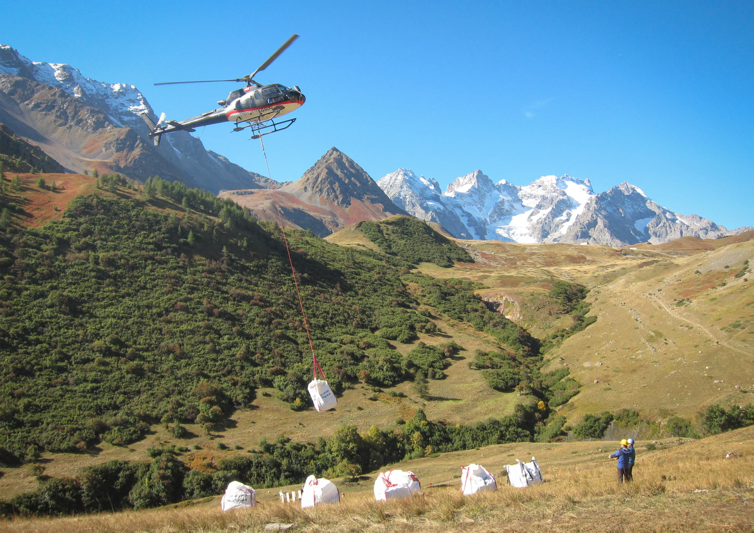 A hunk of earth dangles off a helicopter as two researchers look on.