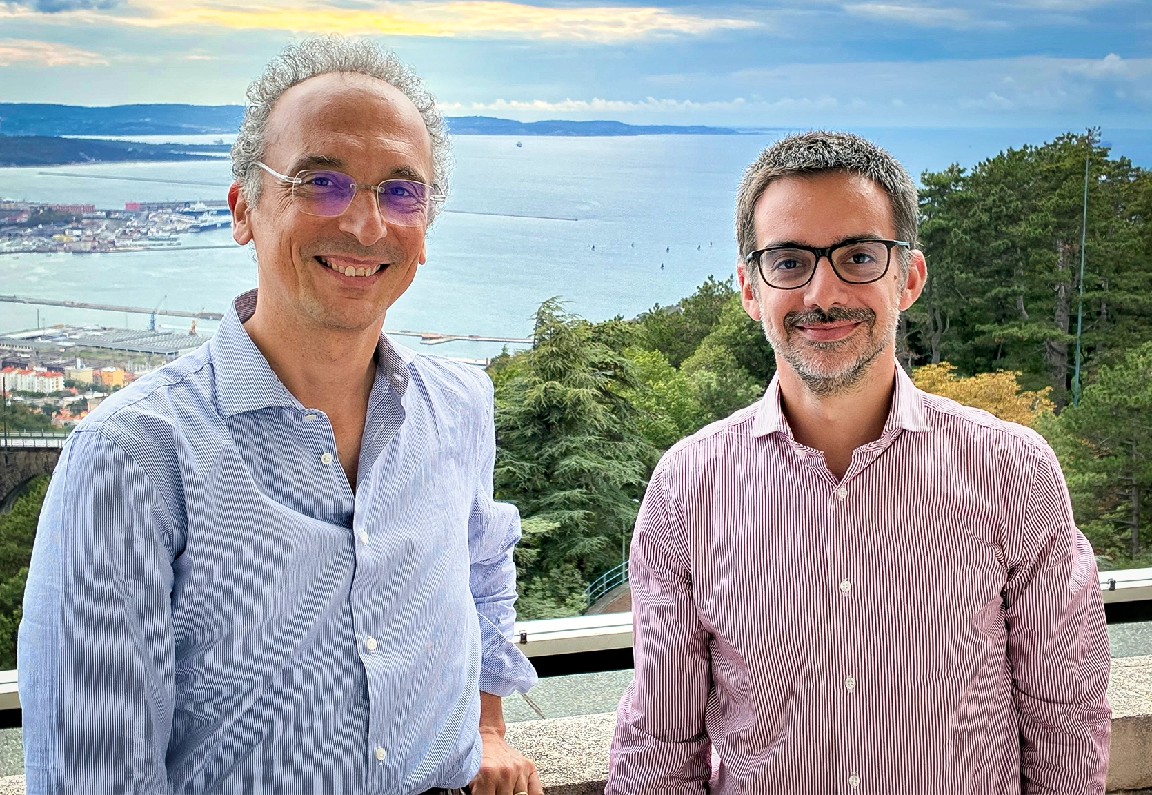 Two men, one wearing a blue shirt and the other a red shirt, standing in front of a view of the Gulf of Trieste.