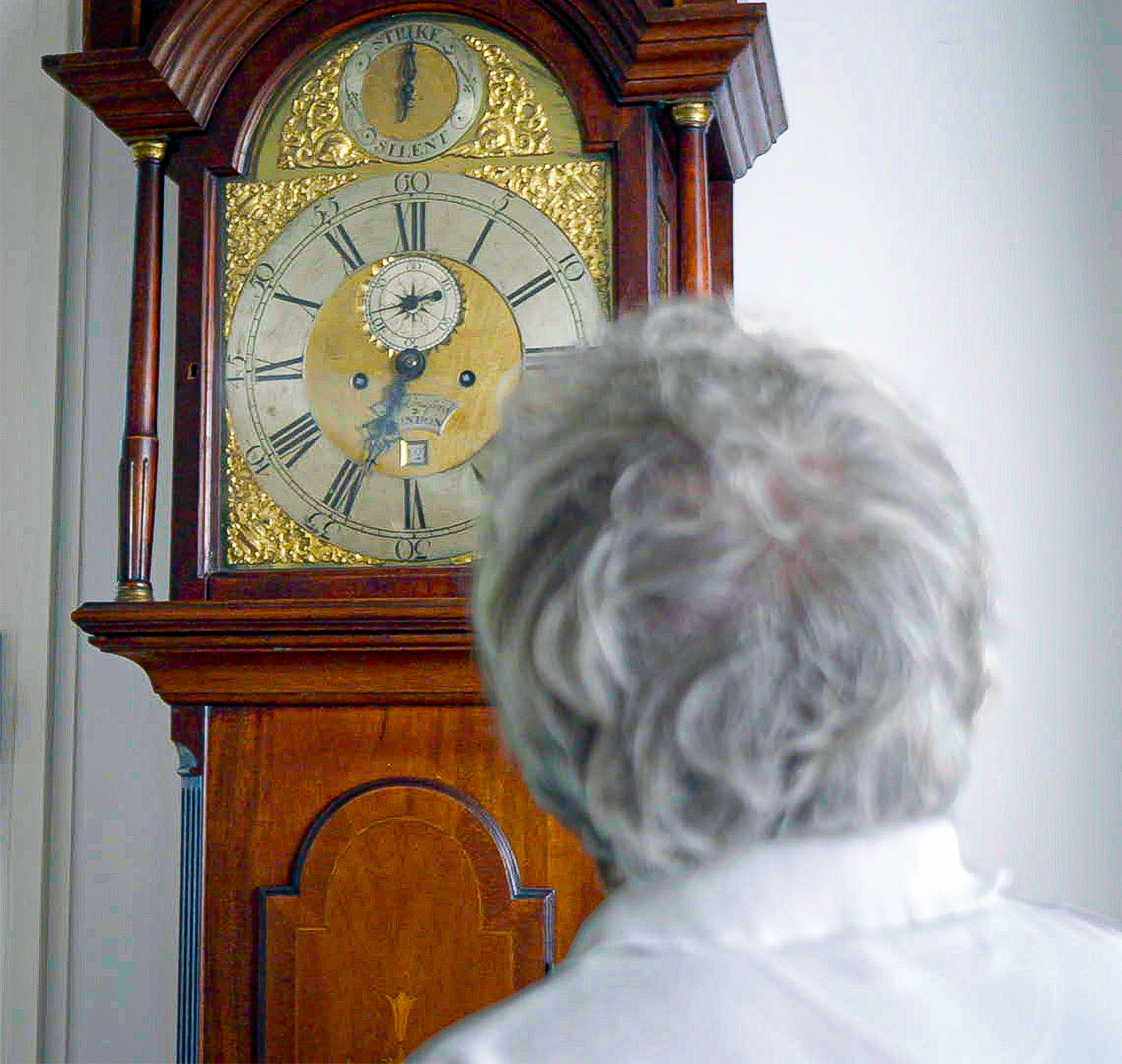 A man is seen from behind, looking up at the face of a grandfather clock.