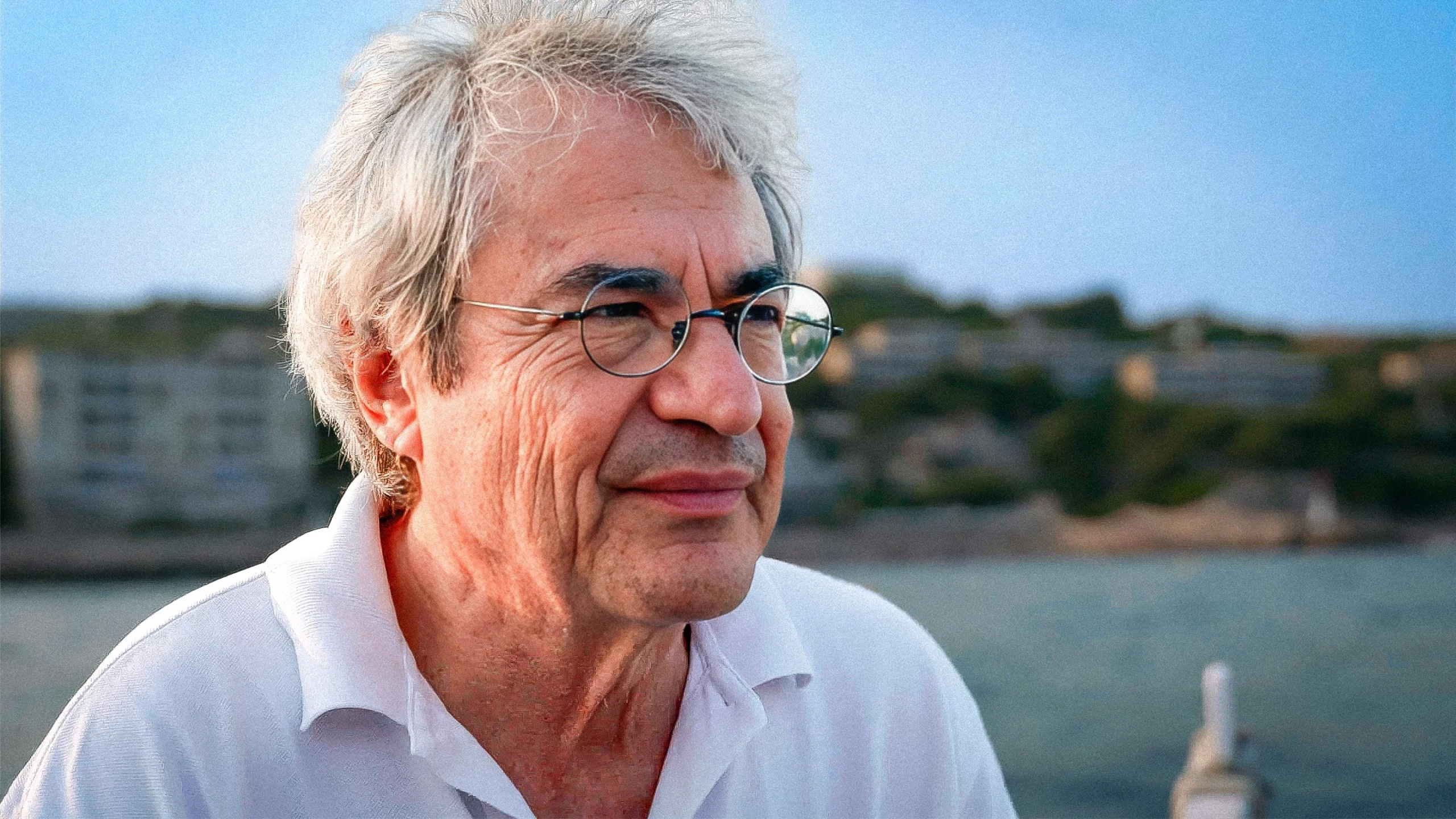 A man with frizzy gray hair and wire-frame glasses peers into the distance in the foreground. He is on a boat on the water with the shoreline visible behind.