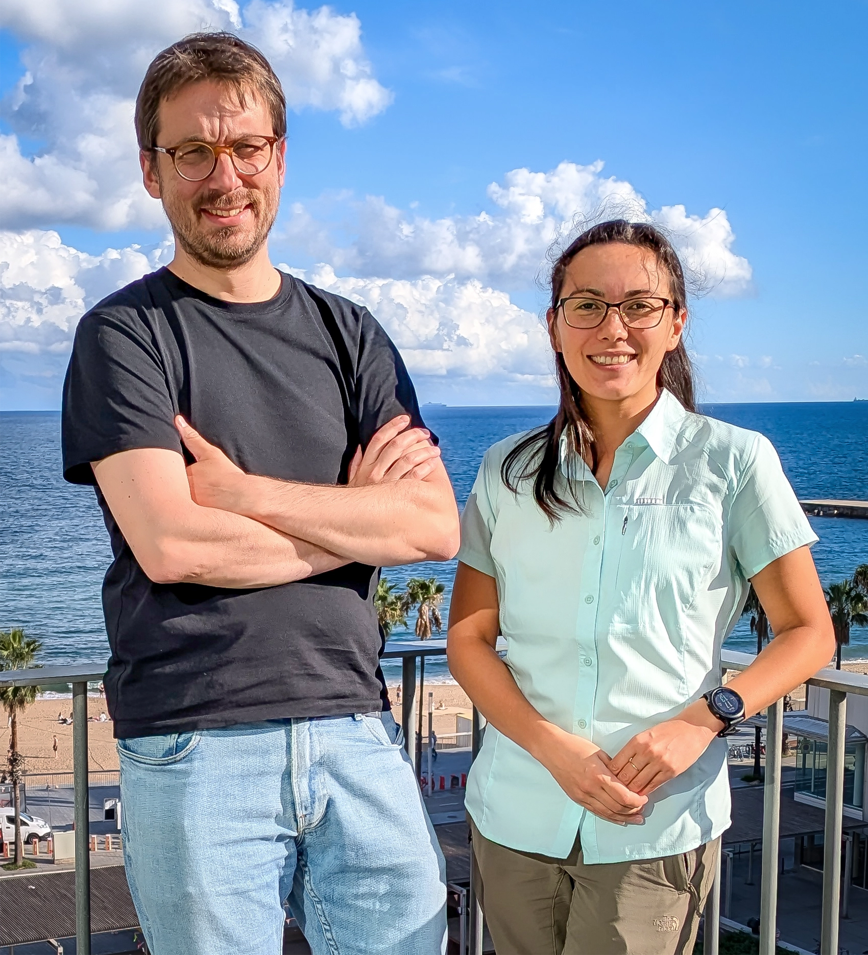 A man and a woman standing together on a balcony with the sea behind them. The man on the left is smiling, wearing a dark T-shirt and jeans, he has short brown hair, glasses and is smiling and has his arms folded. The woman on the right is shorter, also wears glasses and is smiling. She is wearing a light blue-green short-sleeved shirt and light brown pants.