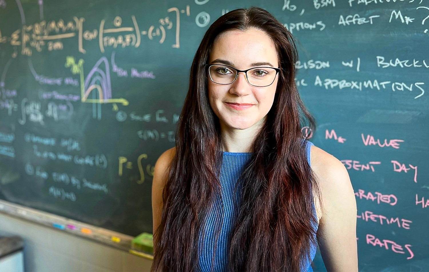 a woman wearing glasses stands in front of a chalkboard