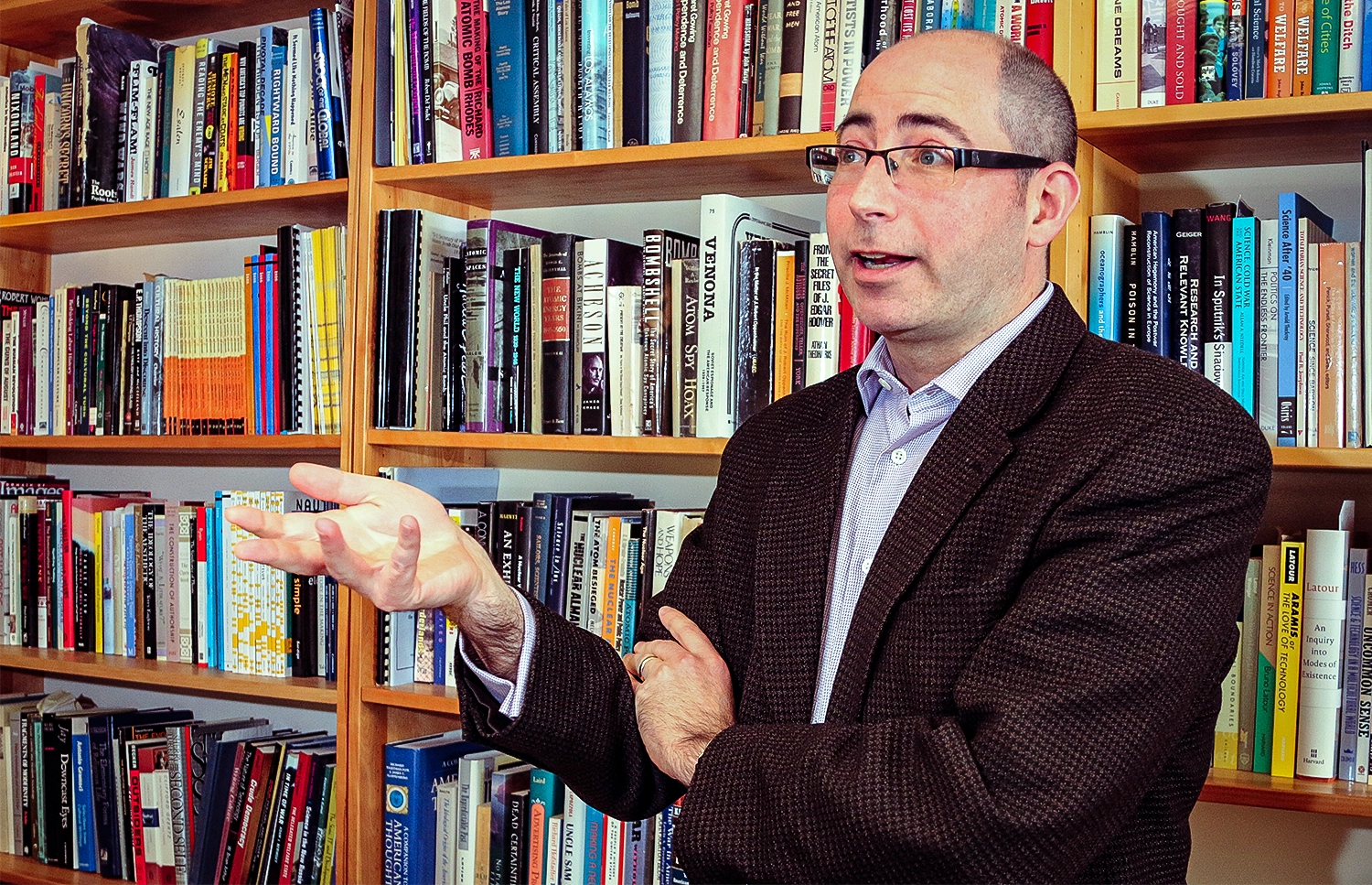 a man wearing glasses stands in front of a bookshelf