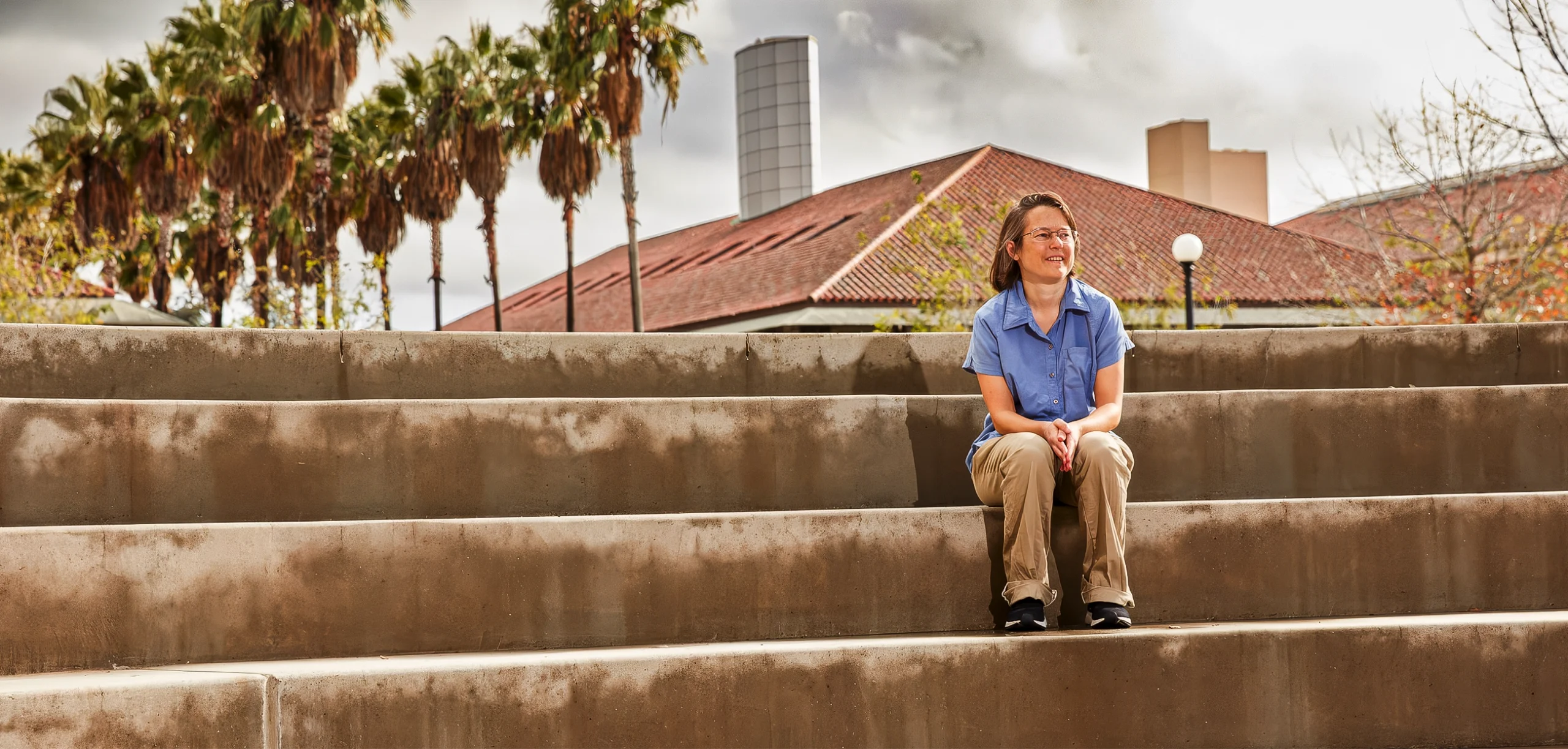 A woman sits on the steps outside near rows of palm trees