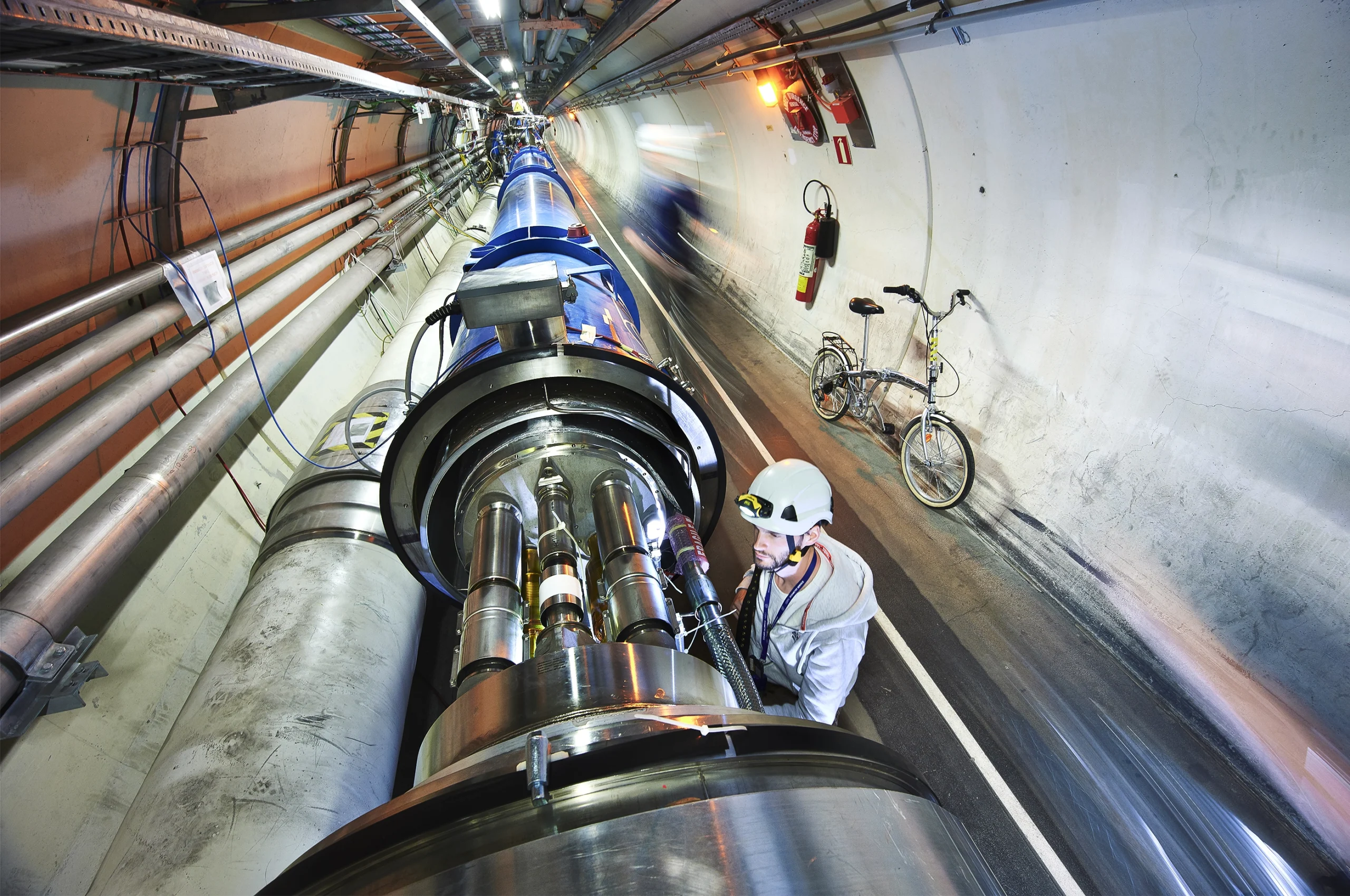 A long scientific tube in a tunnel is examined by a man in a helmet, a bike rests nearby.