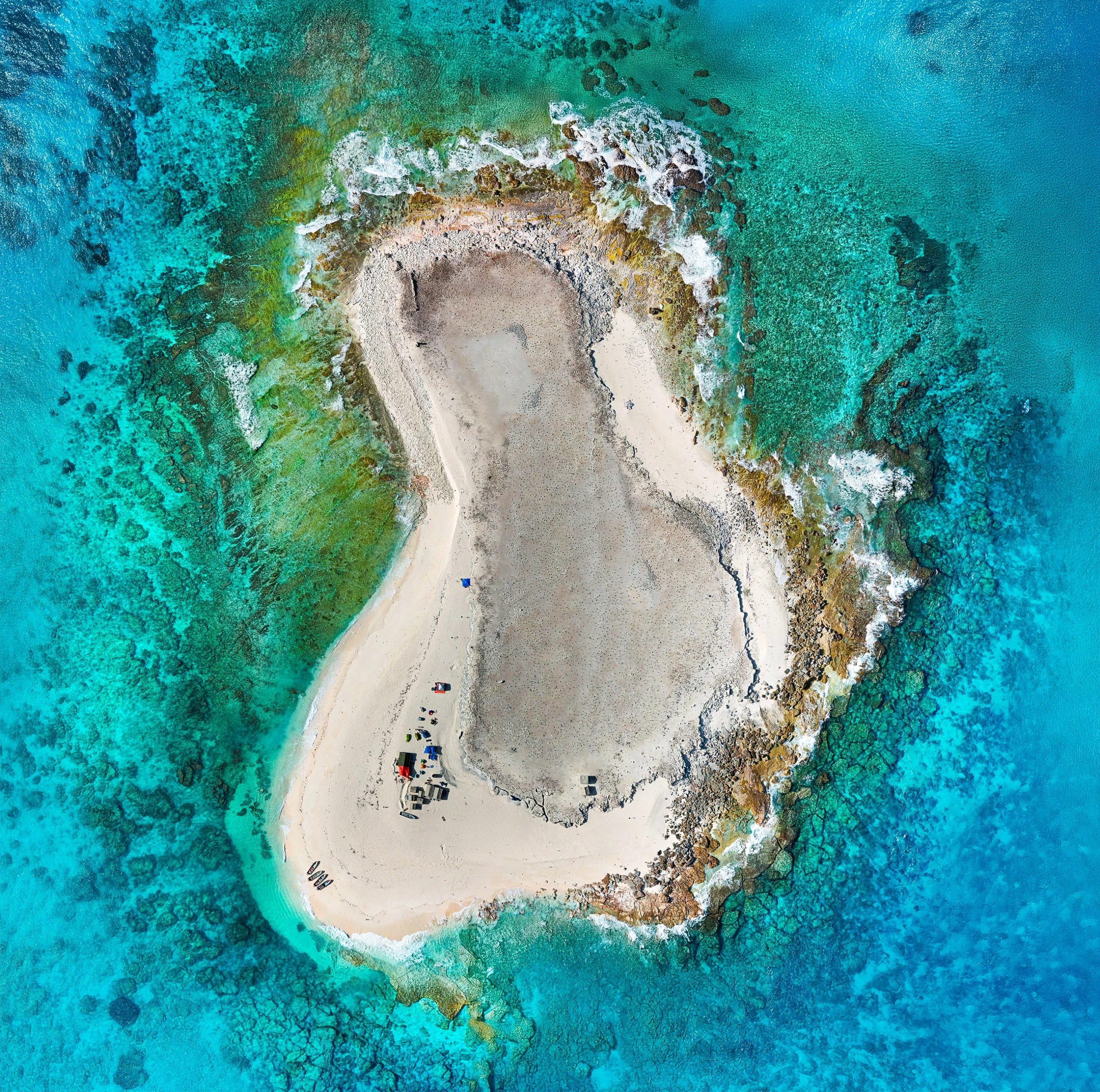 An overhead photo shows a bean-shaped sandy island surrounded by turquoise water. A small cluster of structures emerges from the bottom-left corner.
