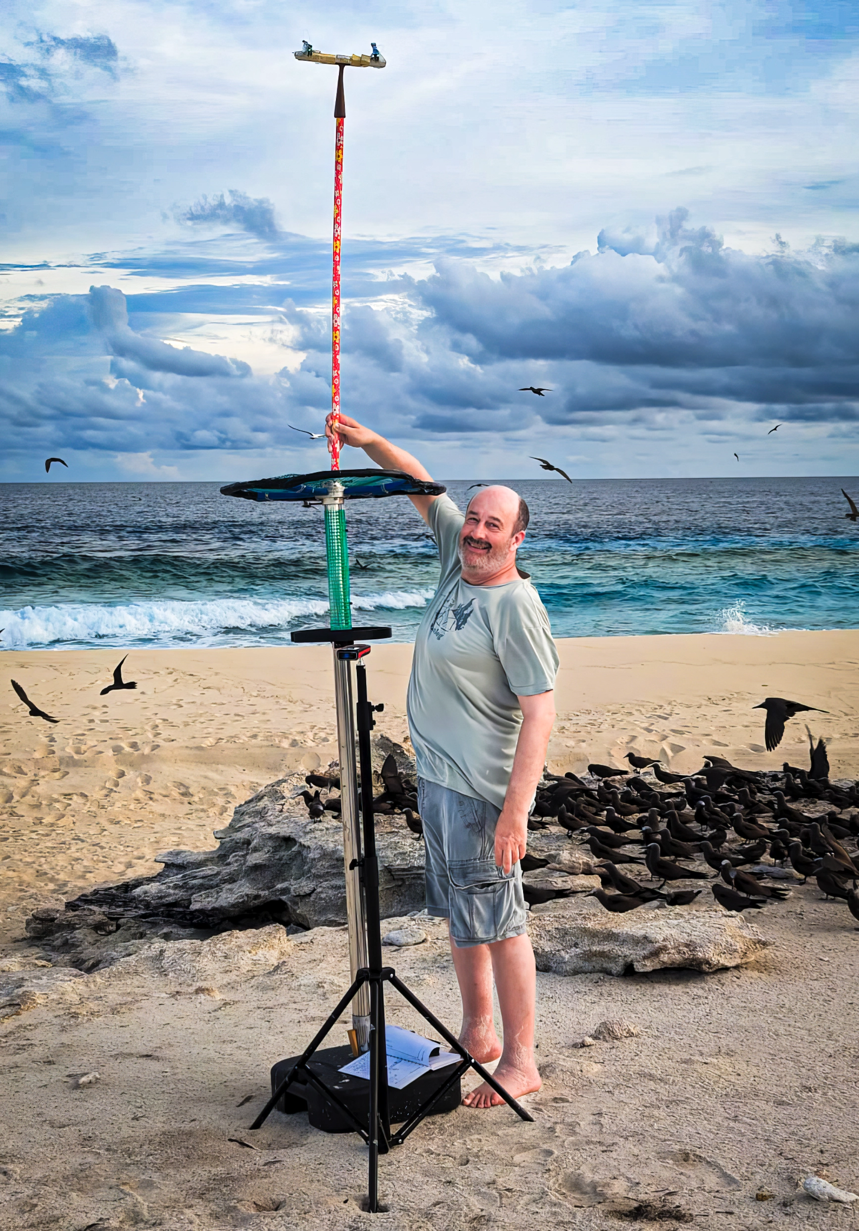 A barefoot Nachum Ulanovsky sets up a tower of scientific equipment on a sandy beach as seabirds crowd behind him.