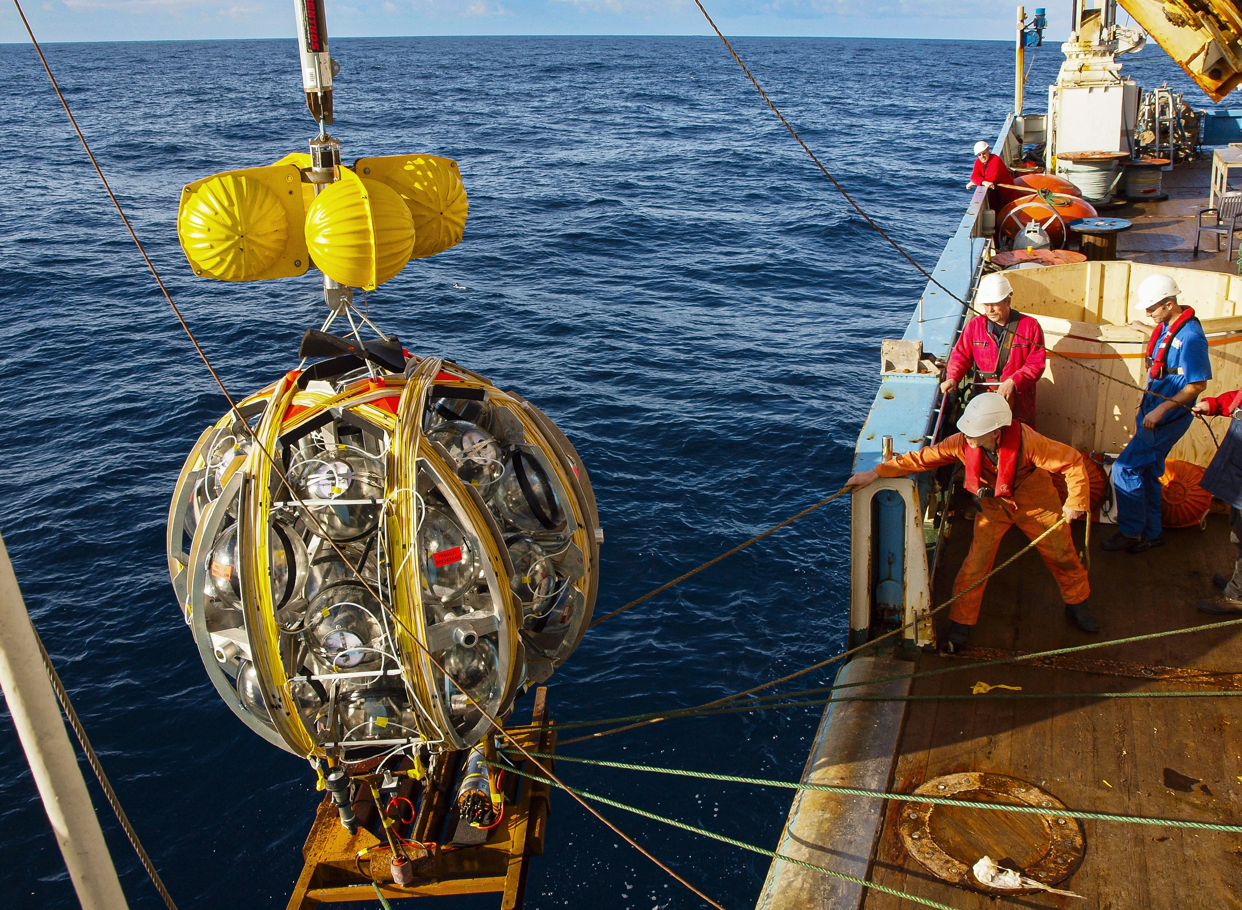 A collection of round detectors, bound together in a spherical frame, is lowered from the side of a ship into the Mediterranean Sea. Workers in brightly colored coveralls and white hard hats guide the frame with ropes.