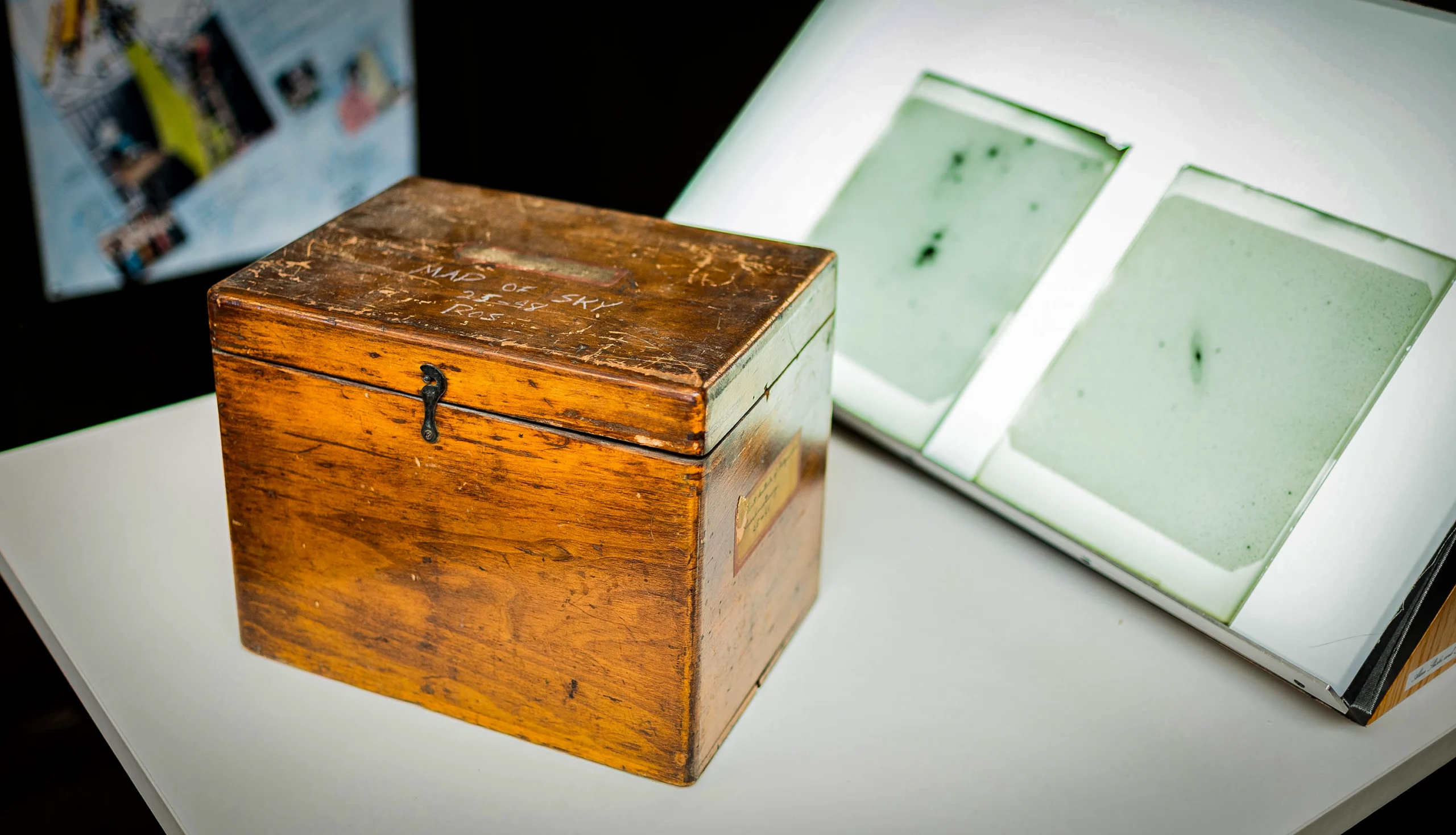 An antique wooden crate sits in front of a tilted light box holding two astronomical negative photographs.