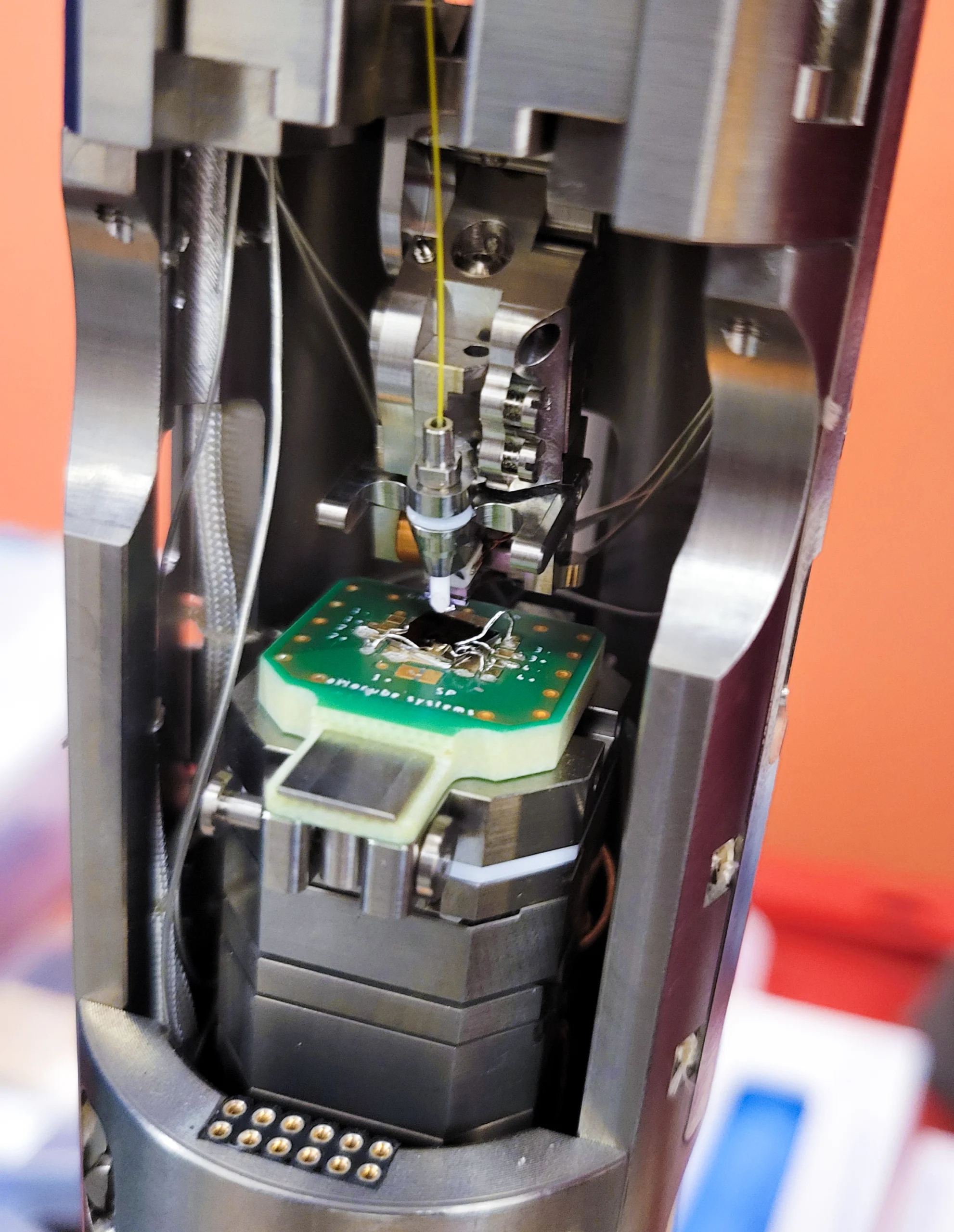 Метална глава лебди изнад платформе унутар лабораторијског уређаја.