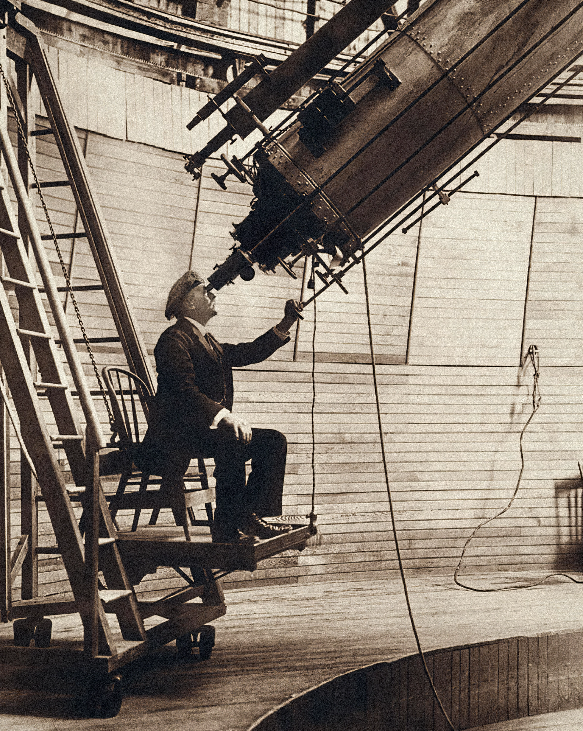 A vintage photo shows a seated man looking in the small eyepiece of a large upward-pointing telescope.