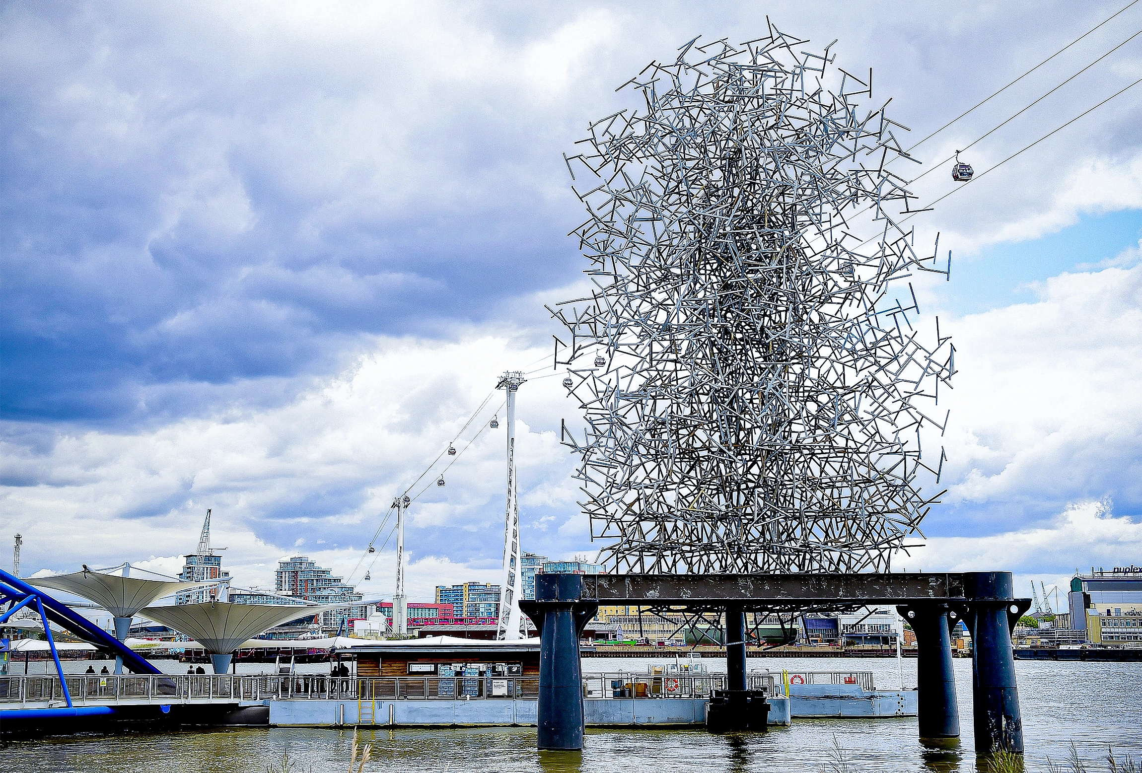 A modern steel sculpture, on a platform over water, looks like a vertical cloud of bars, but has the shape of a figure in the center.