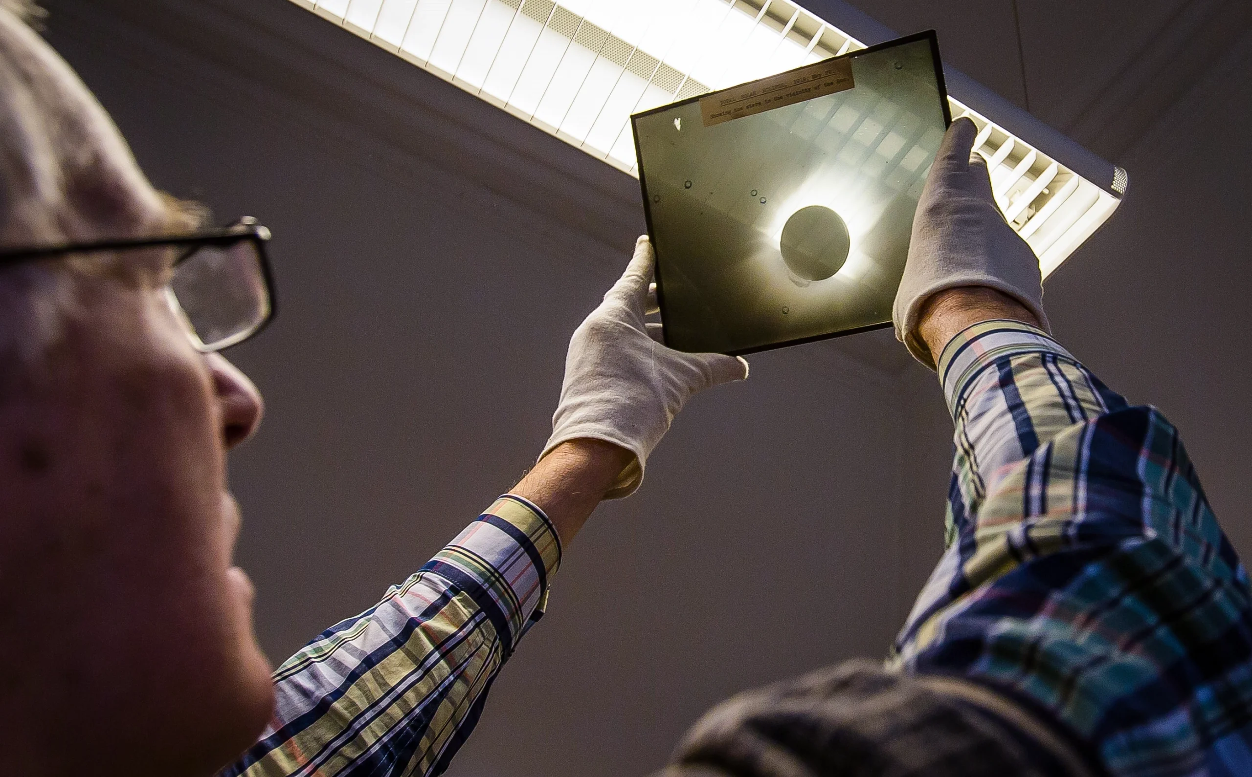 A man holds a photographic plate depicting a solar eclipse up to an overhead light fixture.