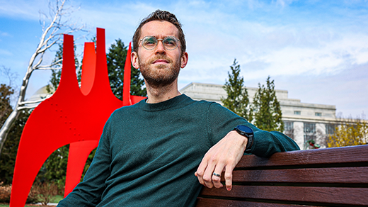 A bespectacled man sitting on a bench in front of a red sculpture