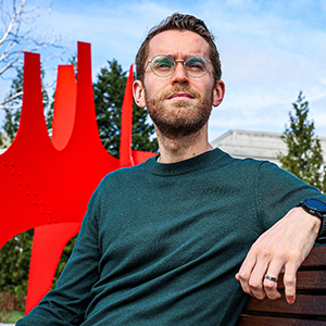 A bespectacled man sitting on a bench in front of a red sculpture
