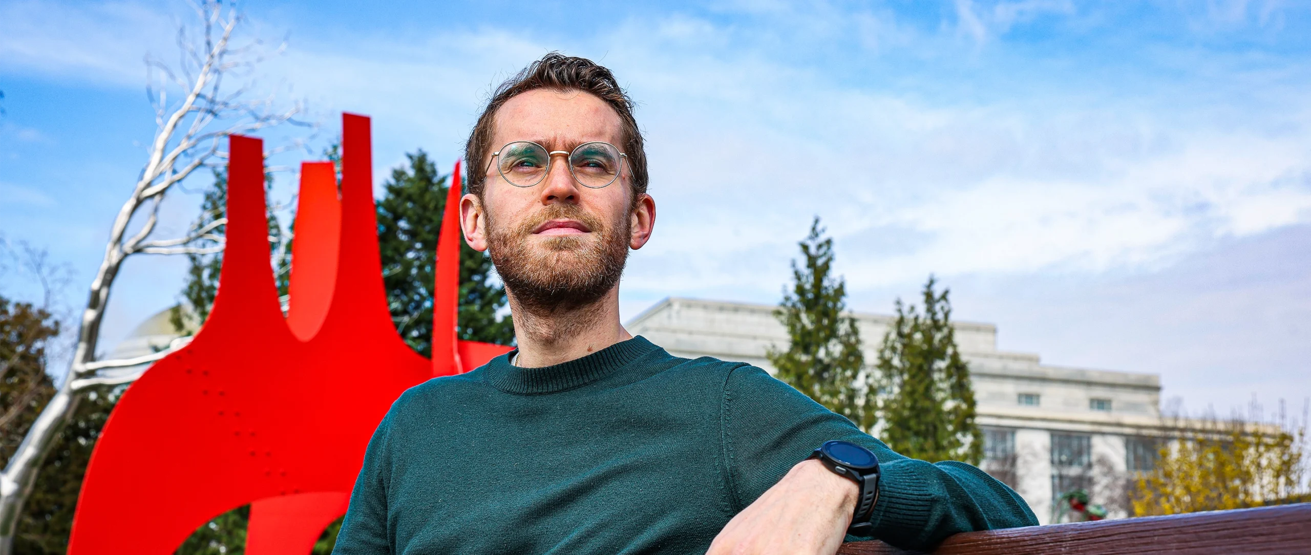 A bespectacled man sitting on a bench in front of a red sculpture