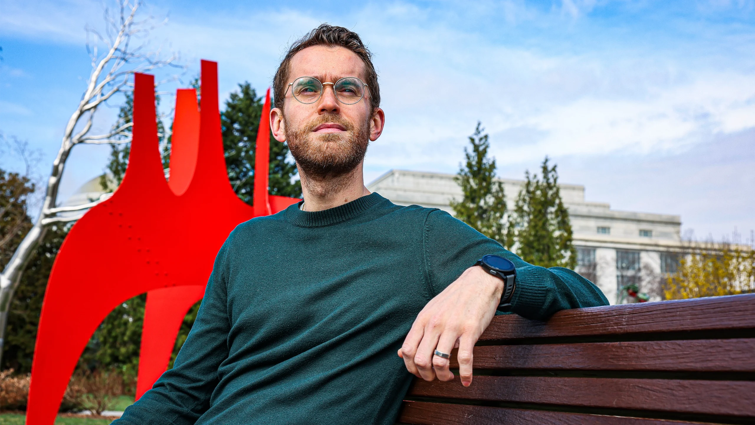 A bespectacled man sitting on a bench in front of a red sculpture