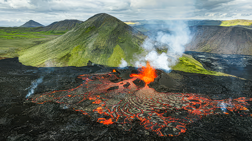 A mountainous landscape is divided in two, with the area closer to the viewer covered in black rock and glowing flows of lava and the area farther from the viewer looking verdant and unaffected by the ongoing eruption.