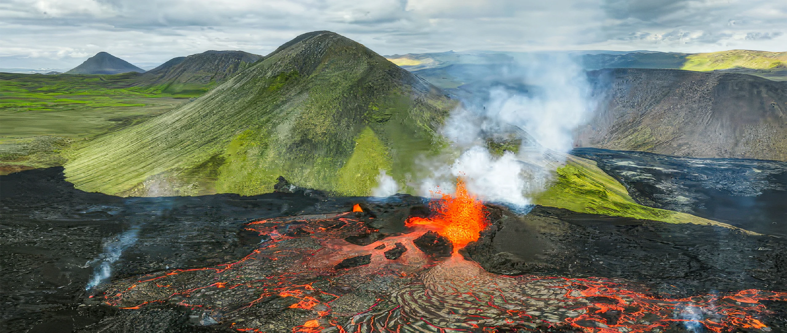 A mountainous landscape is divided in two, with the area closer to the viewer covered in black rock and glowing flows of lava and the area farther from the viewer looking verdant and unaffected by the ongoing eruption.