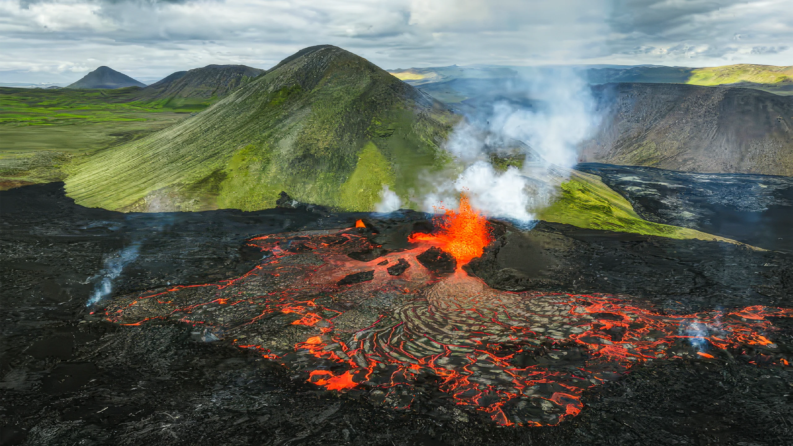 A mountainous landscape is divided in two, with the area closer to the viewer covered in black rock and glowing flows of lava and the area farther from the viewer looking verdant and unaffected by the ongoing eruption.